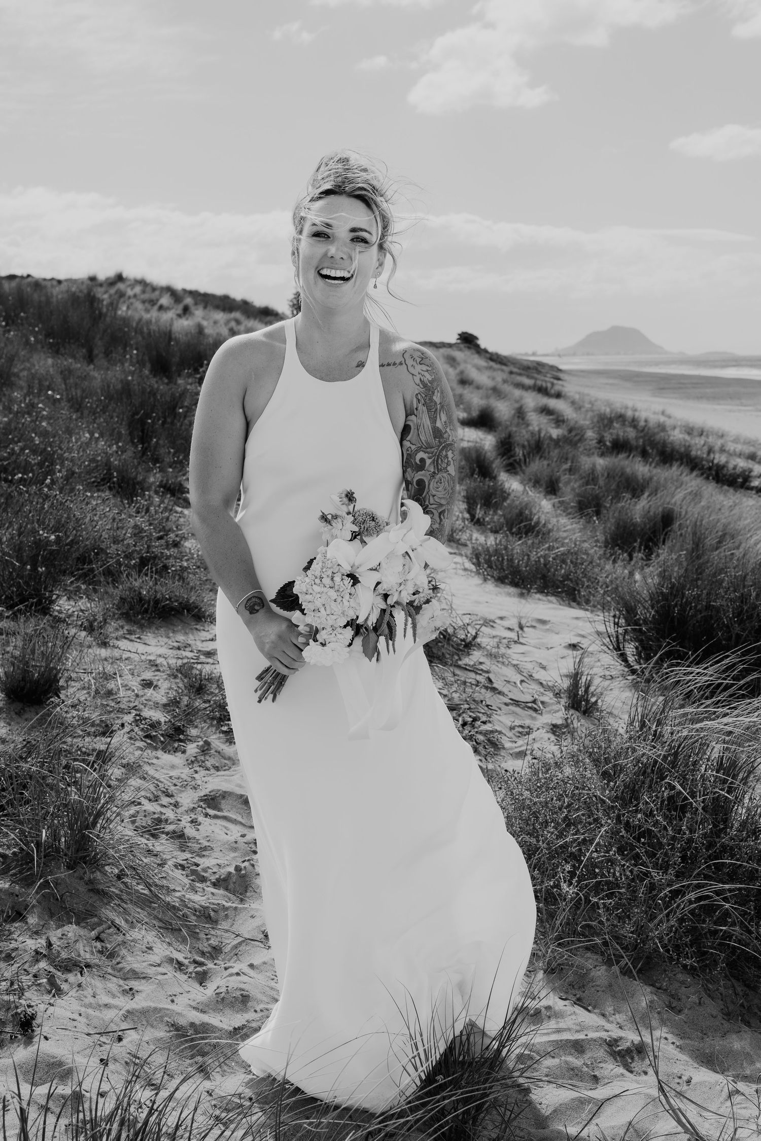 Smiling bride in white halter gown holds floral bouquet on sandy beach dunes in black and white photo.
