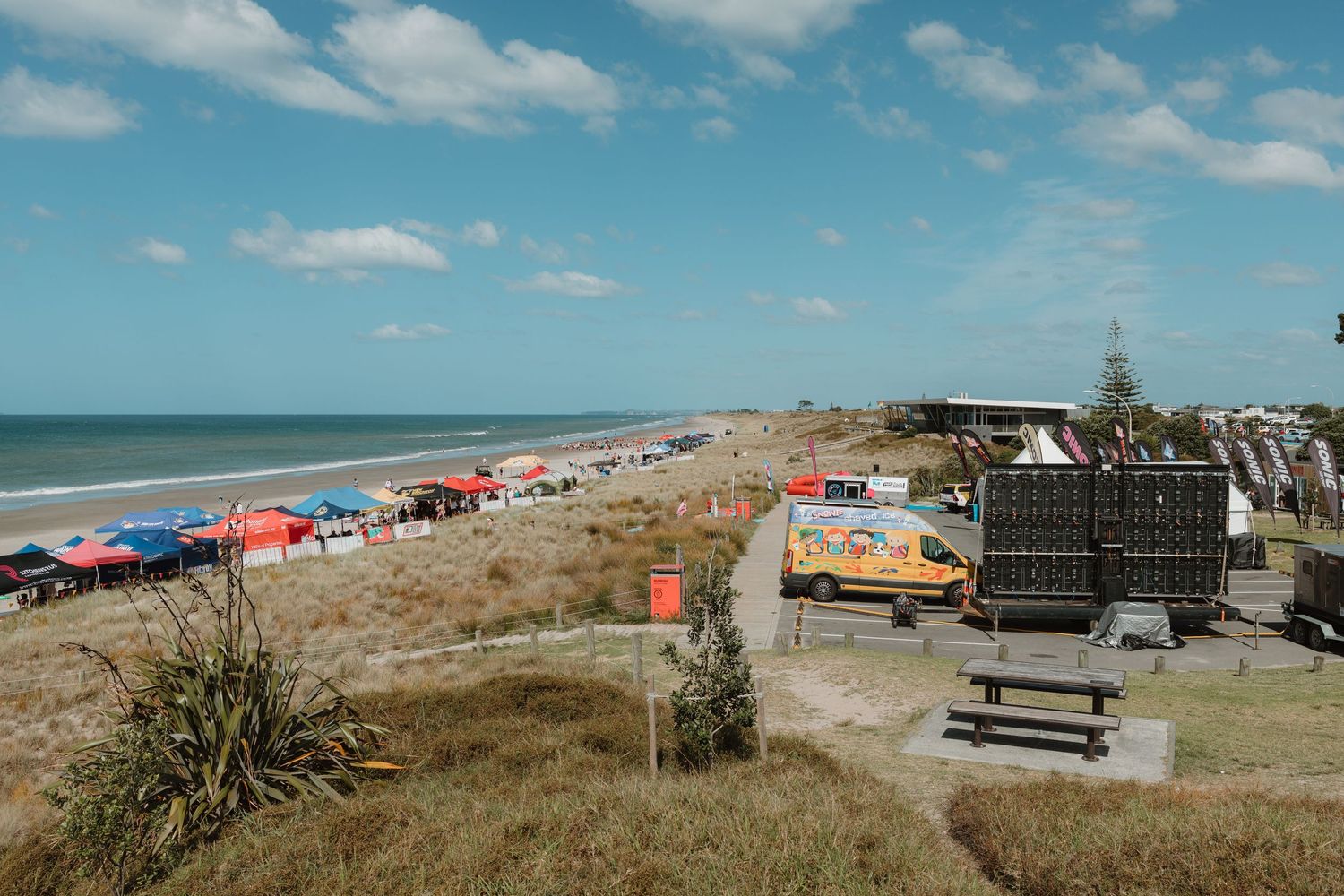 Aerial view of a busy beach with colorful umbrellas, food trucks, and vendors along the sandy New Zealand coastline.