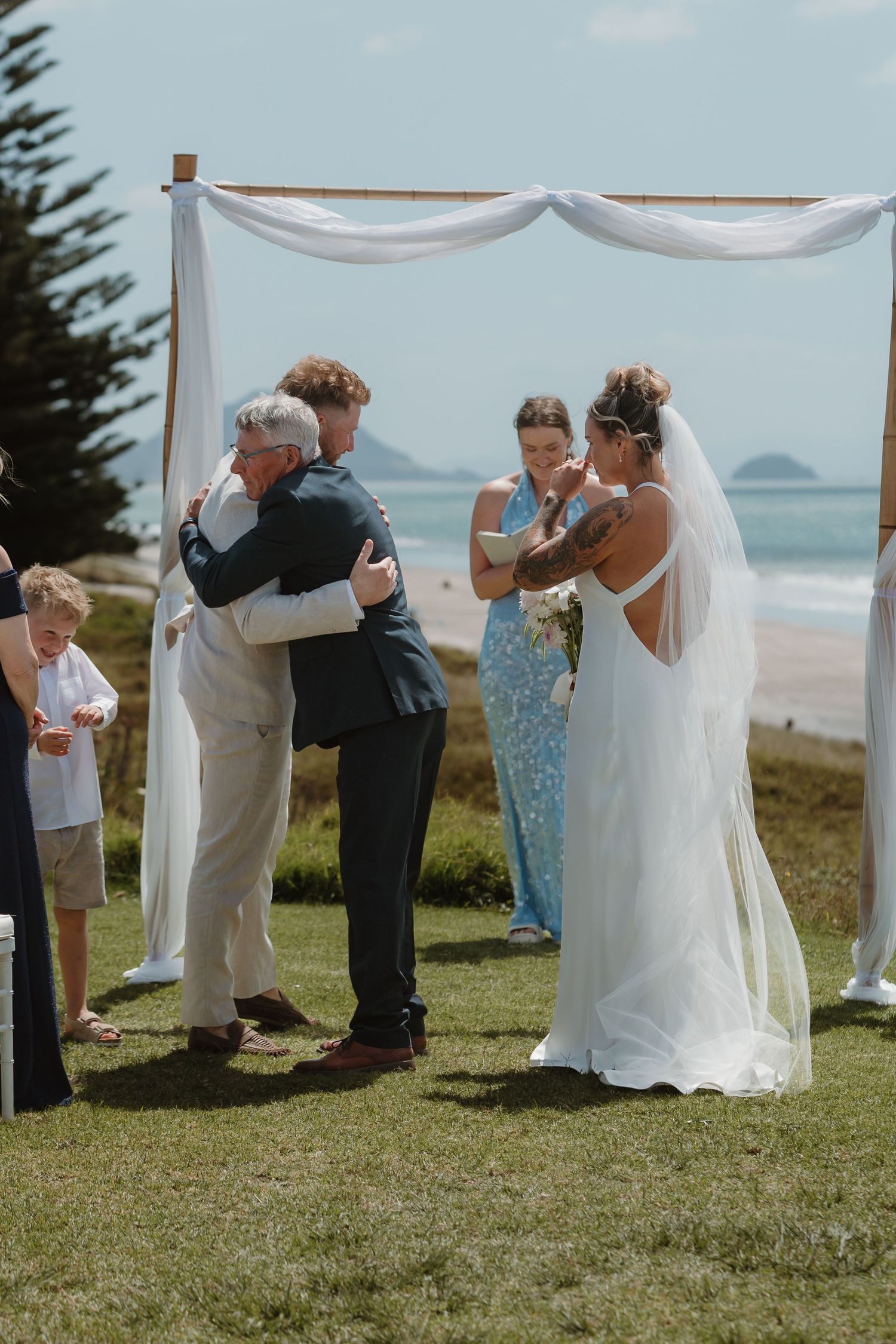 Bride and groom share emotional hug with father at outdoor beach wedding ceremony under white draped arch.