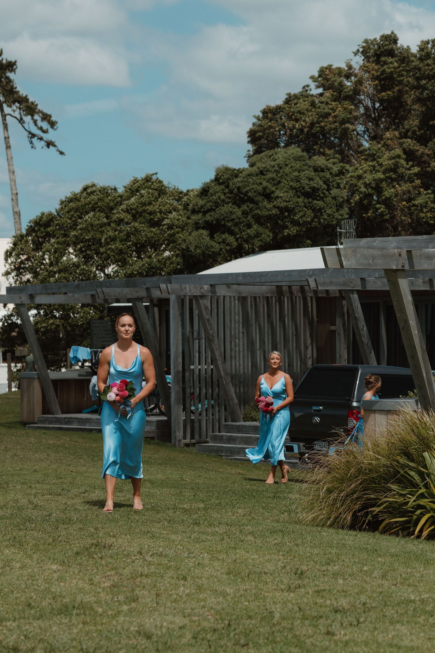 Two bridesmaids in teal satin dresses carrying pink bouquets walk barefoot across green lawn at outdoor wedding venue.