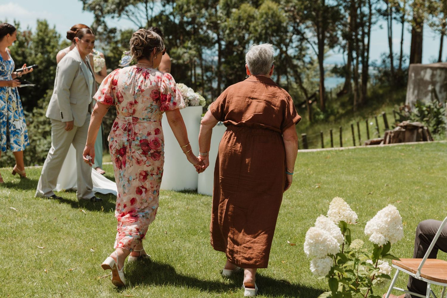 Two women holding hands walk across a sunny green lawn at an outdoor wedding ceremony.