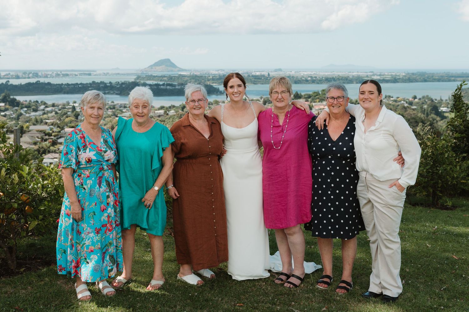 Bride in white gown poses with six women in colorful dresses on green hillside with scenic coastal views.
