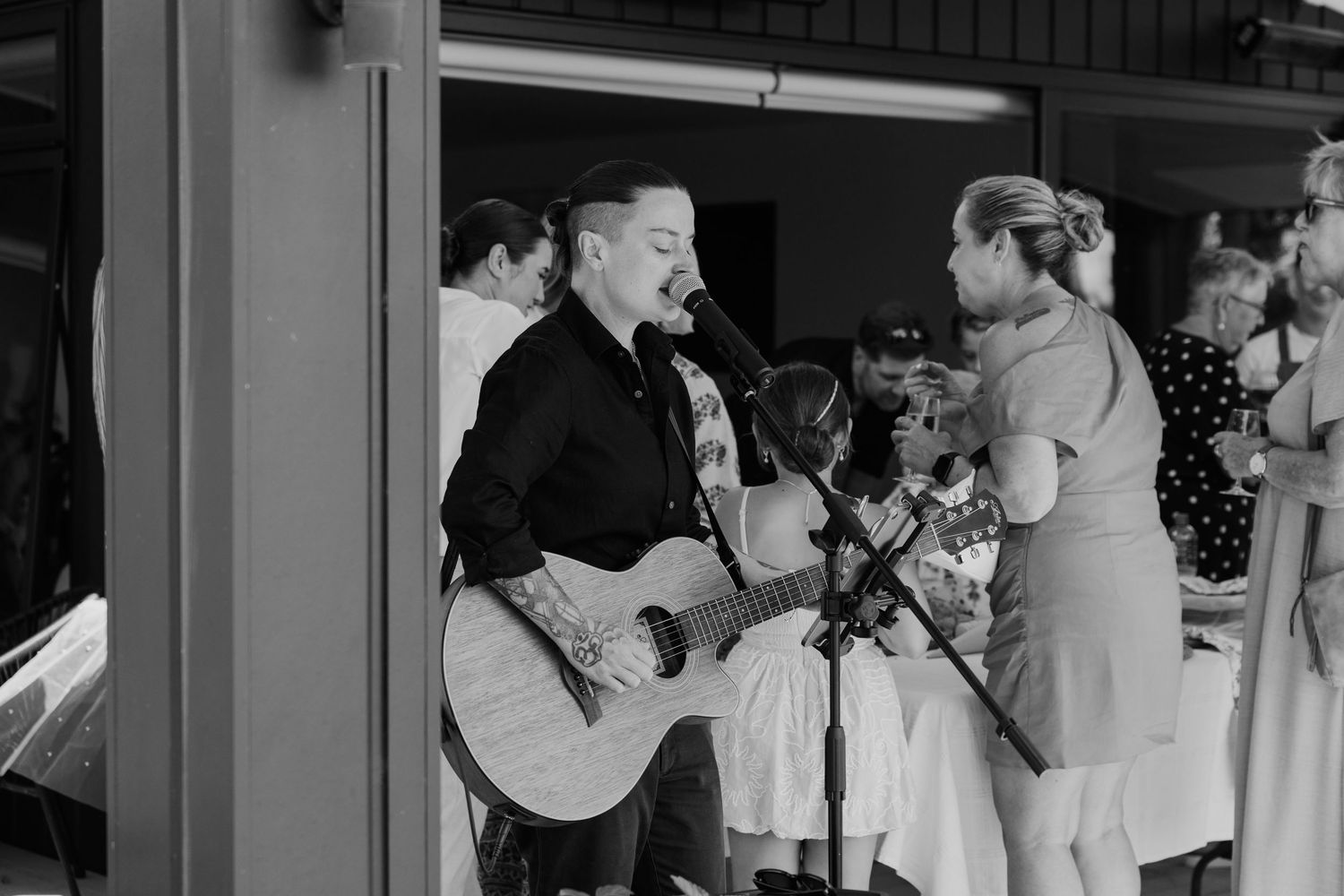 Black and white photo of a tattooed musician playing acoustic guitar and singing into a microphone at an outdoor event.