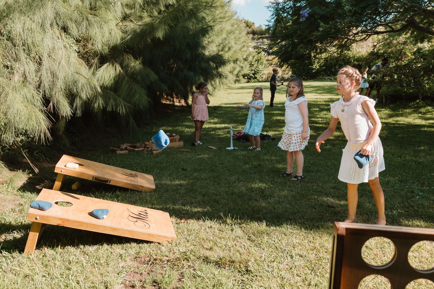 Children playing cornhole and washer toss lawn games outdoors on green grass surrounded by trees.