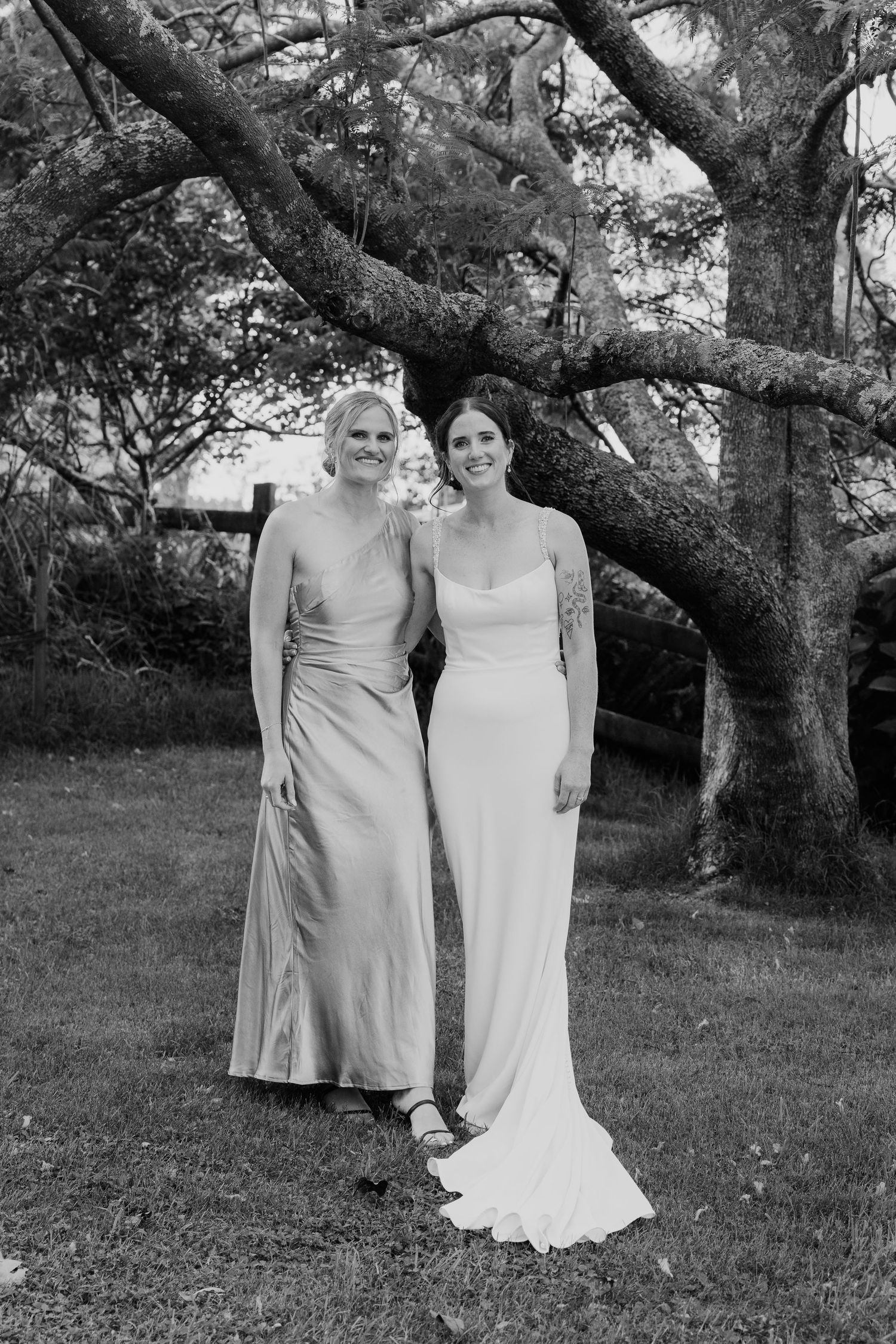 Black and white photo of bride in white gown and bridesmaid in satin dress posing under large tree outdoors.