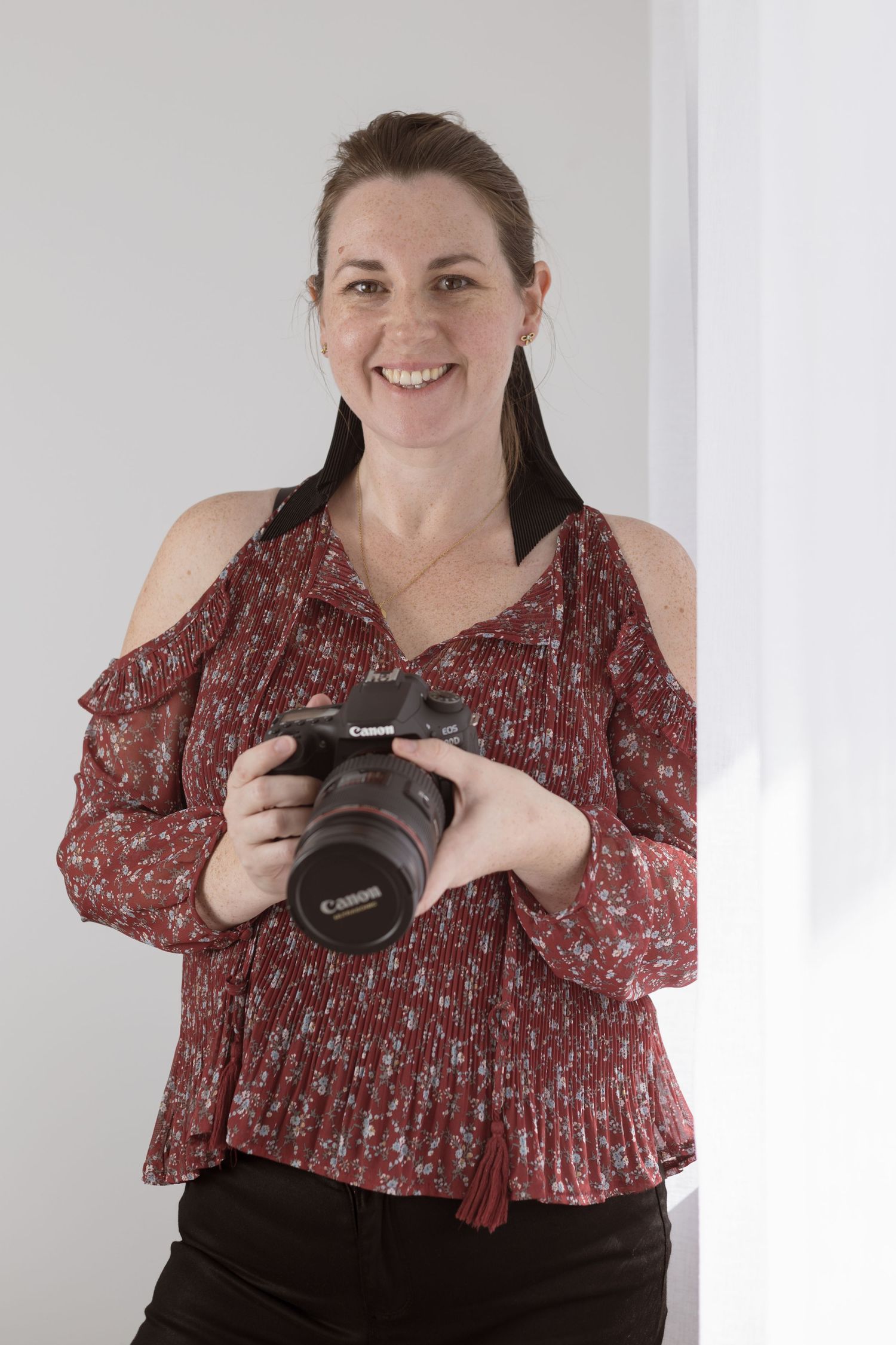 Someone holds a professional DSLR camera while wearing a red floral print top and black pants against a white wall.