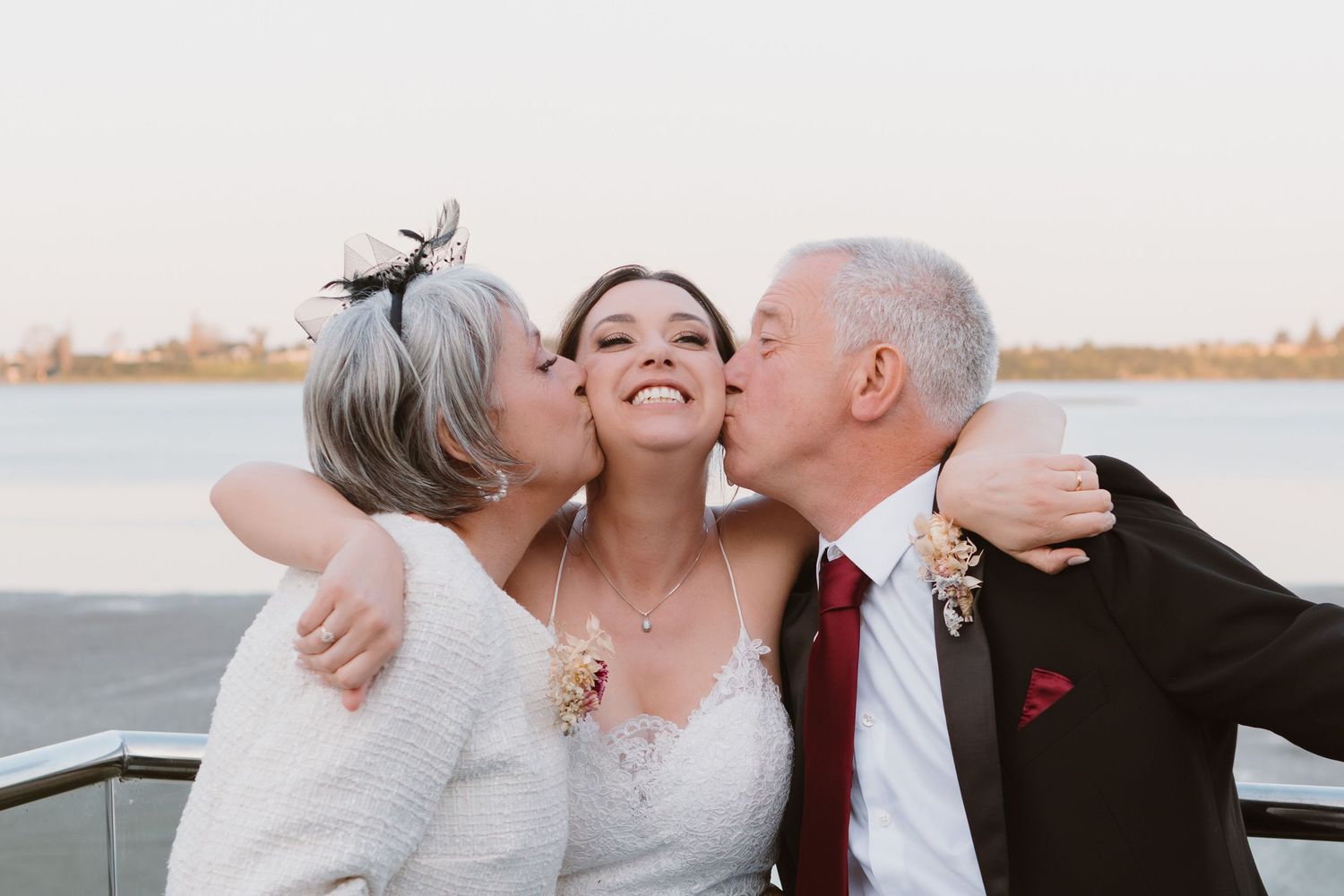 Three people share a joyful embrace and kisses outdoors by the water on a bright day.