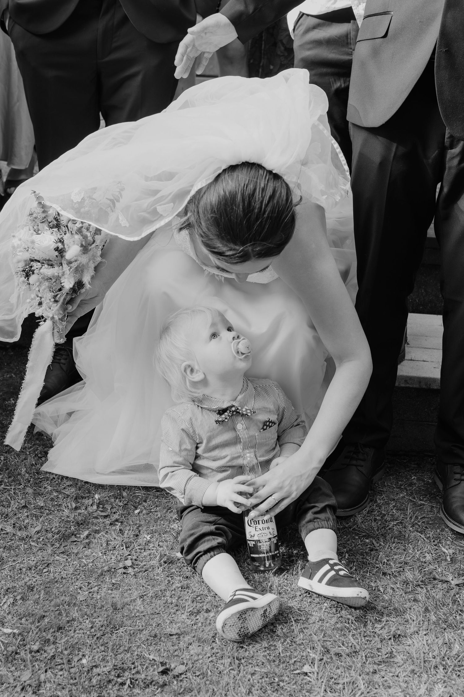 A tender moment between a bride in white dress bending down to comfort a small child at an outdoor wedding event.
