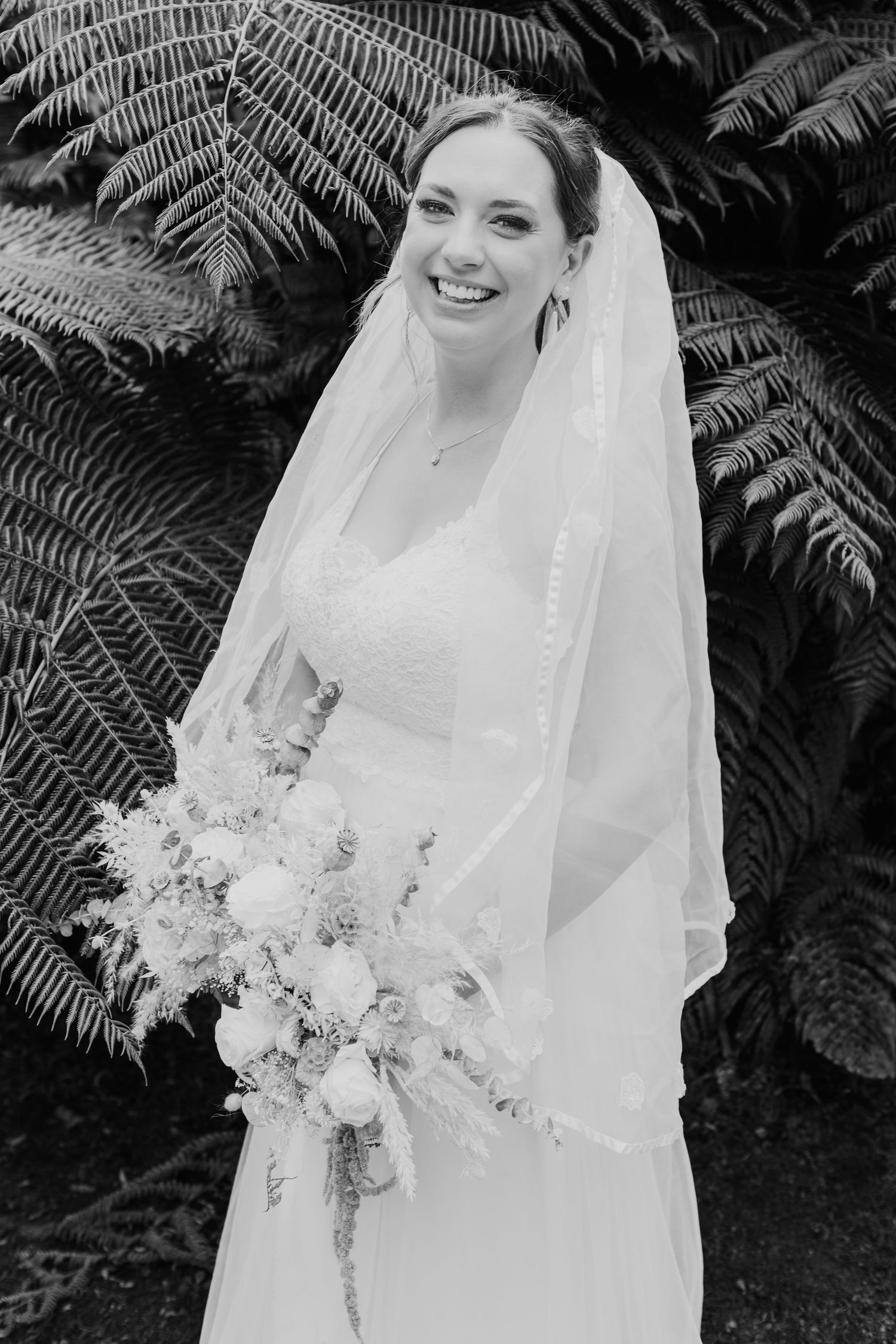 Black and white bridal portrait featuring a flowing veil and elegant bouquet against a backdrop of ferns.