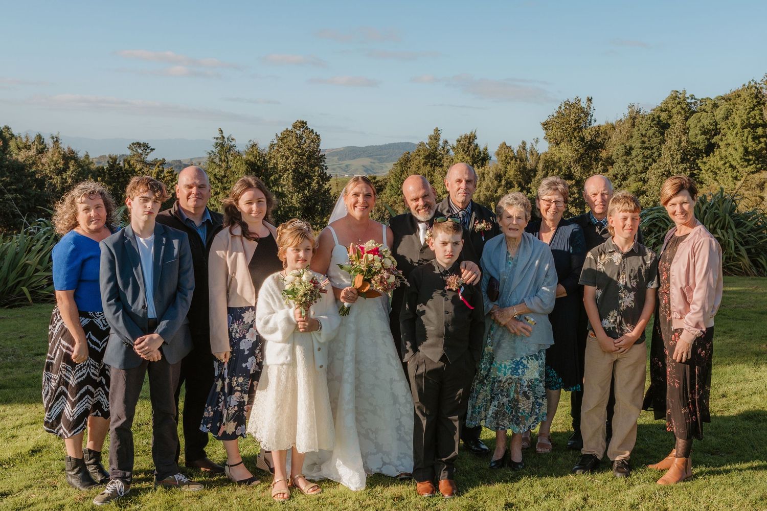 Group standing outdoors on green hilltop with trees and mountains in background during a sunny autumn wedding celebration.