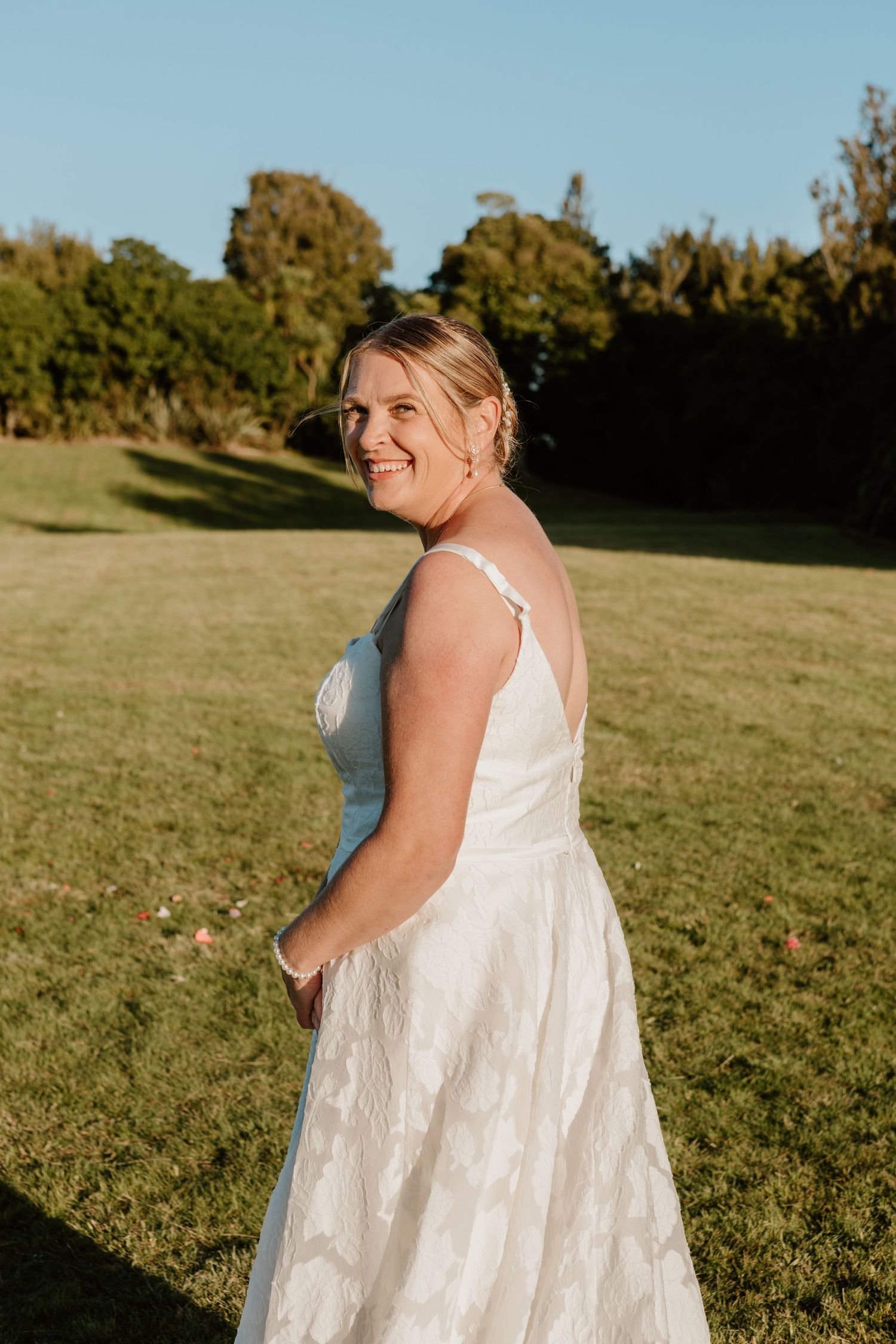 A person in a flowing white lace wedding dress stands on a grassy lawn during golden hour with trees in background.