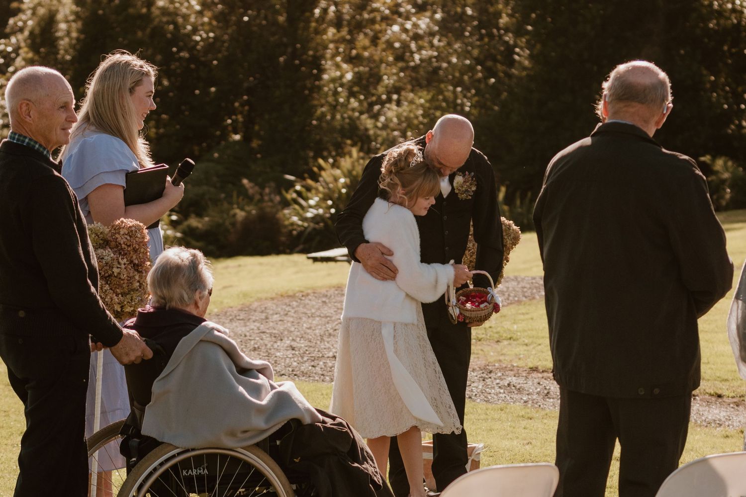 People gather outdoors in formal attire for a sunny wedding celebration in a garden setting.