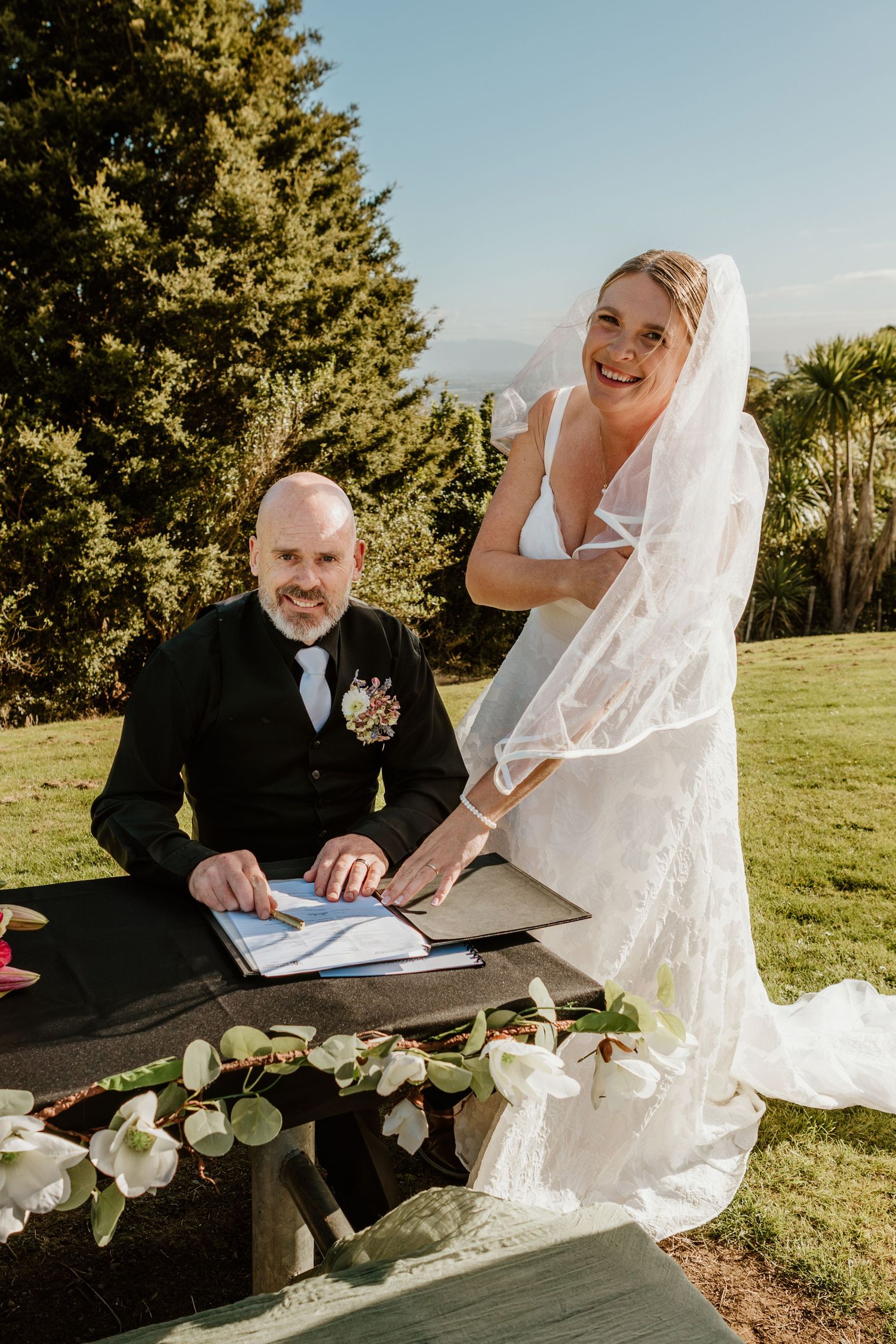 A couple signs their marriage license outdoors during a sunset wedding ceremony with white floral decorations.