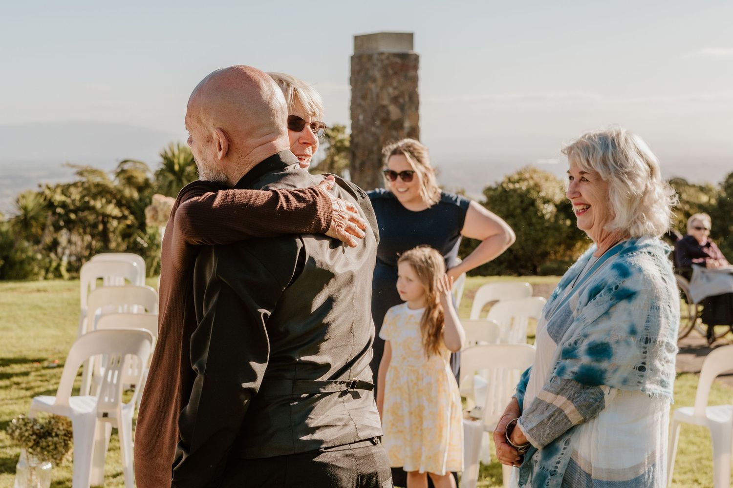 Emotional outdoor celebration moment at sunset with guests gathered on a grassy hilltop near a stone monument.