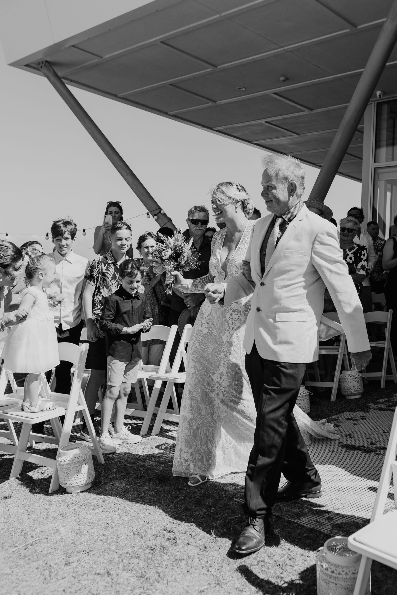 A black and white wedding ceremony photo showing a couple walking down the aisle while guests look on at an outdoor venue.