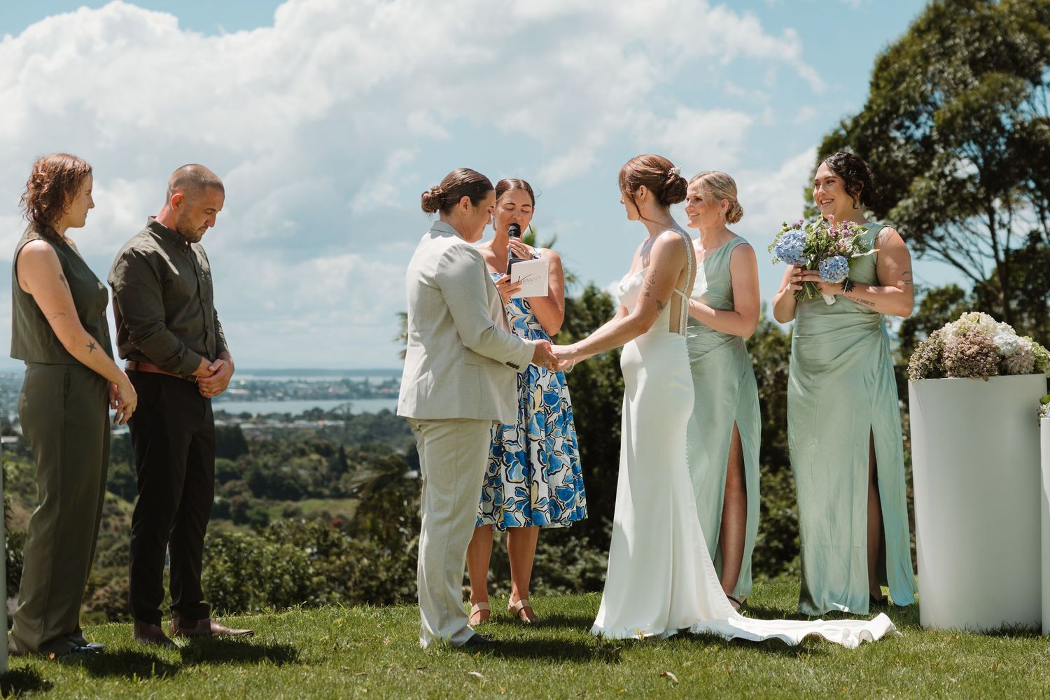 Outdoor wedding ceremony on a hilltop with bride and groom holding hands, bridesmaids in sage green, overlooking a scenic bay.
