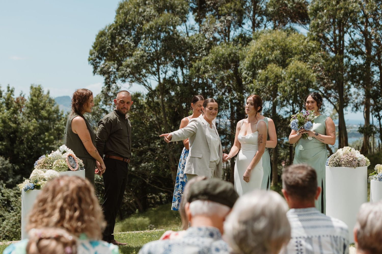 Outdoor wedding ceremony with couple at altar, bridesmaids in mint dresses, and seated guests surrounded by lush green trees.
