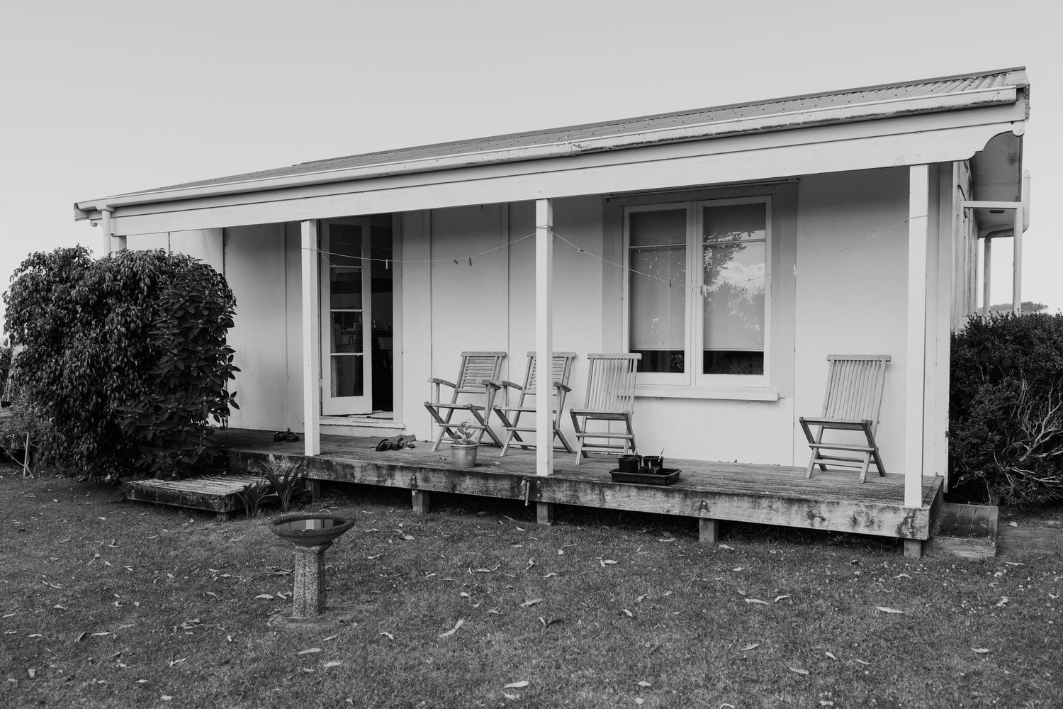 Black and white photo of a quaint cottage with a wooden veranda featuring two relaxing chairs on a grassy lawn.