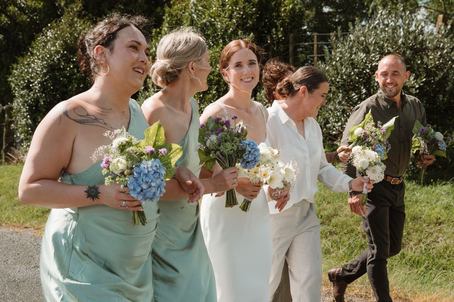 Bride and bridesmaids in mint dresses walk outdoors holding blue and white bouquets, laughing joyfully on a sunny wedding day.