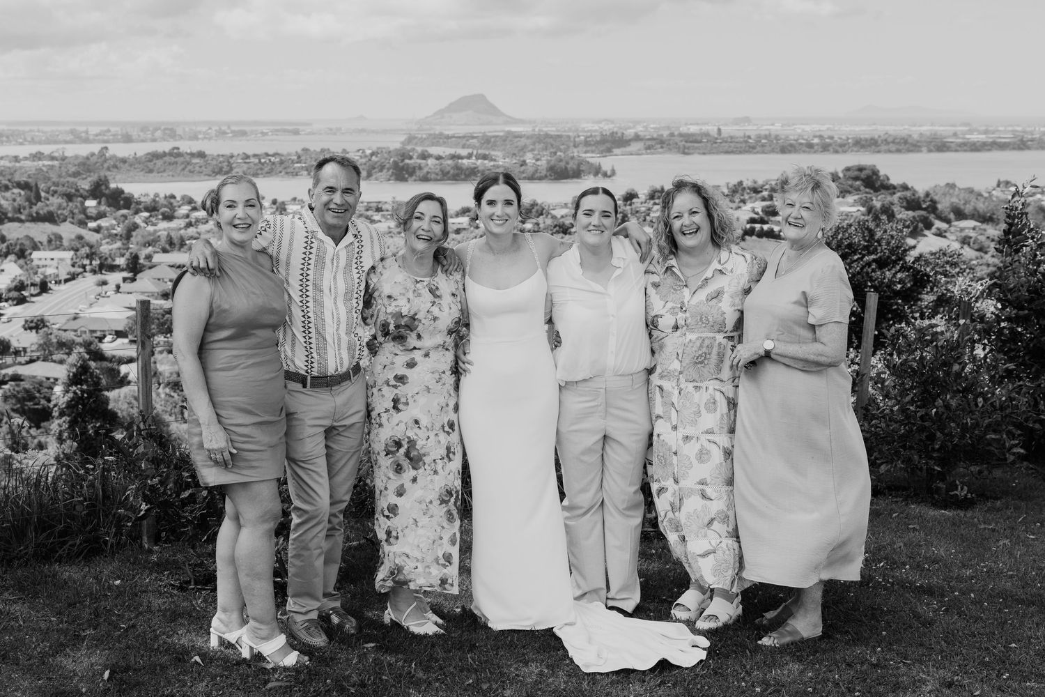 Black and white wedding photo of bride with six guests smiling outdoors with scenic coastal landscape in background.