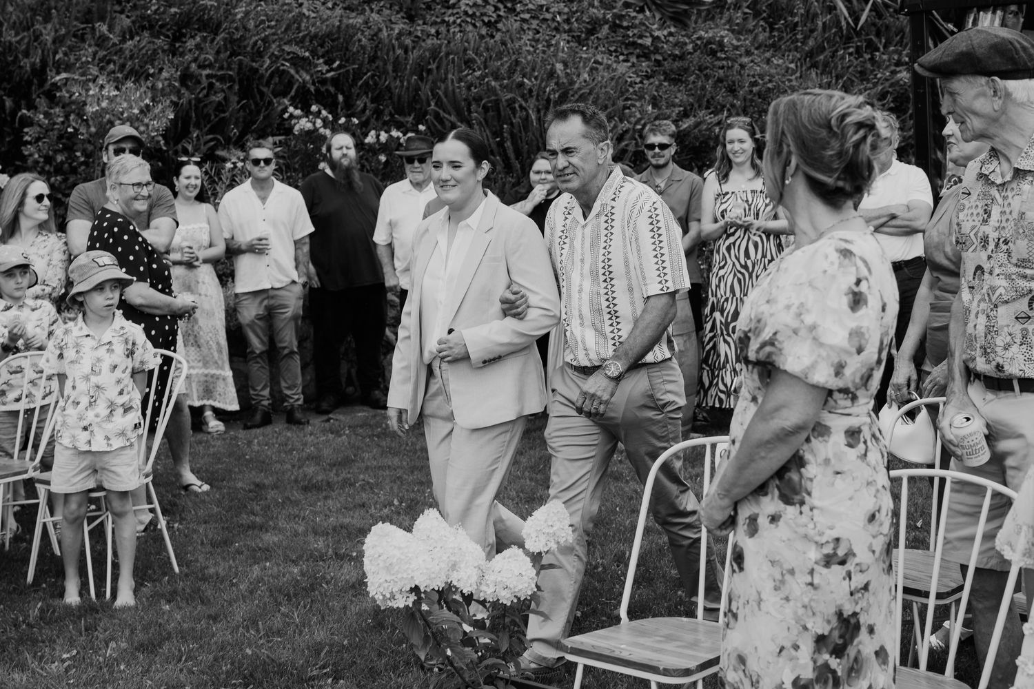 Black and white outdoor wedding ceremony with guests seated on chairs along a garden aisle as couple walks through.