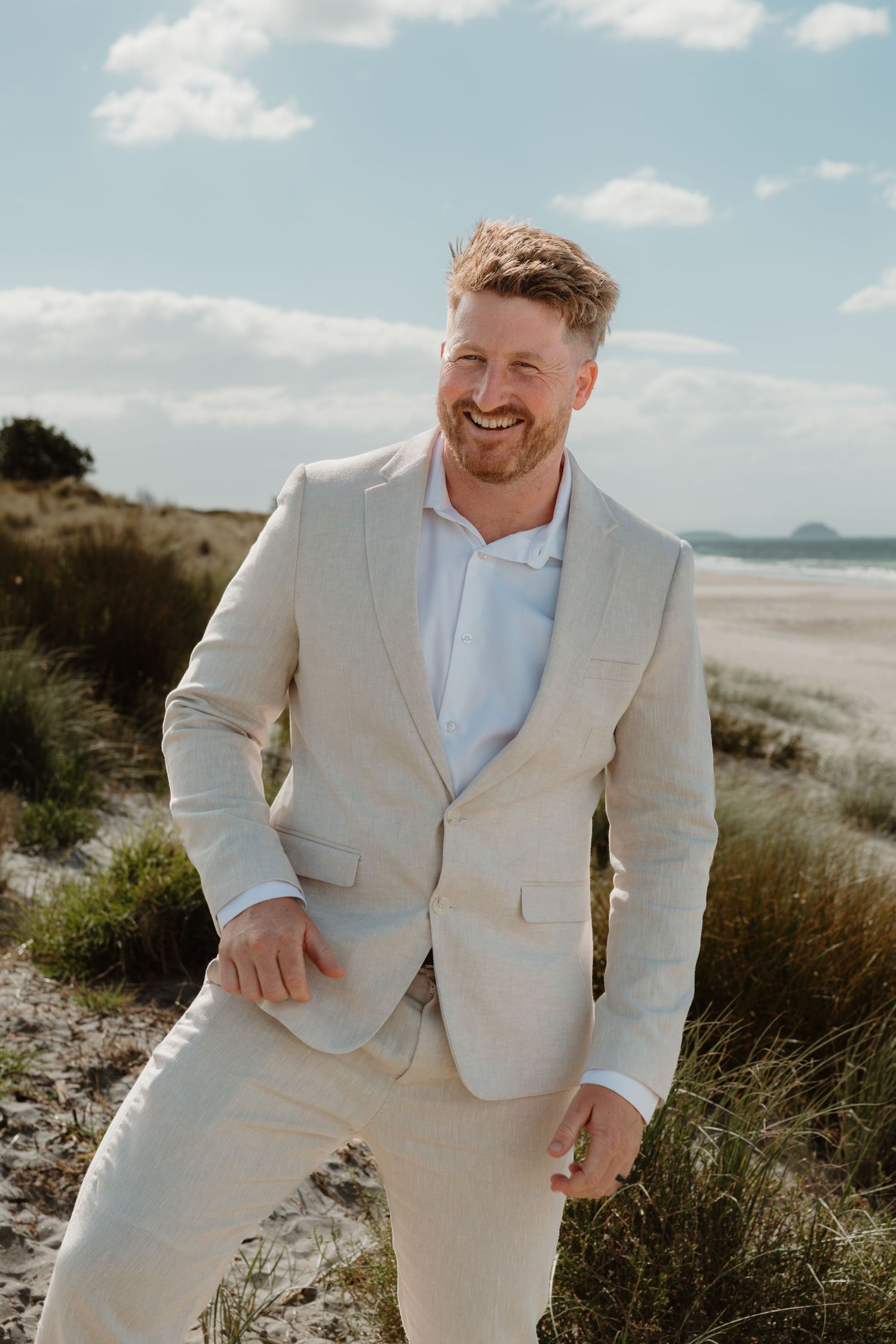 Smiling man in beige linen suit standing on sandy beach with grass dunes and blue sky backdrop.