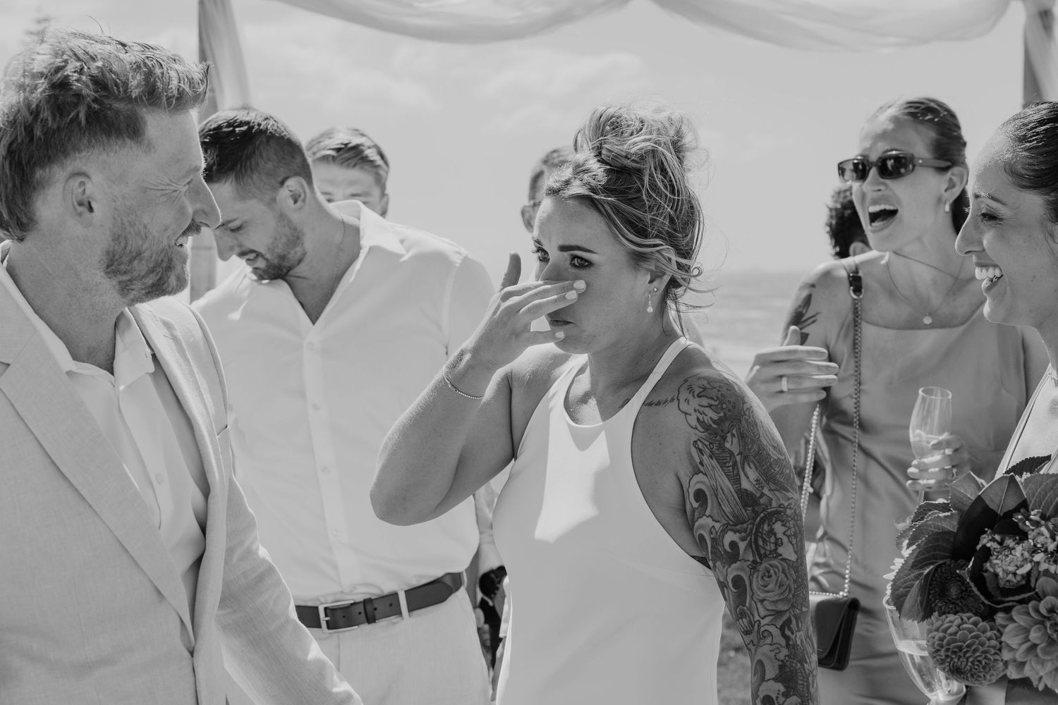 Black and white photo of wedding guests laughing and celebrating outdoors, woman with tattoos wiping tears of joy.