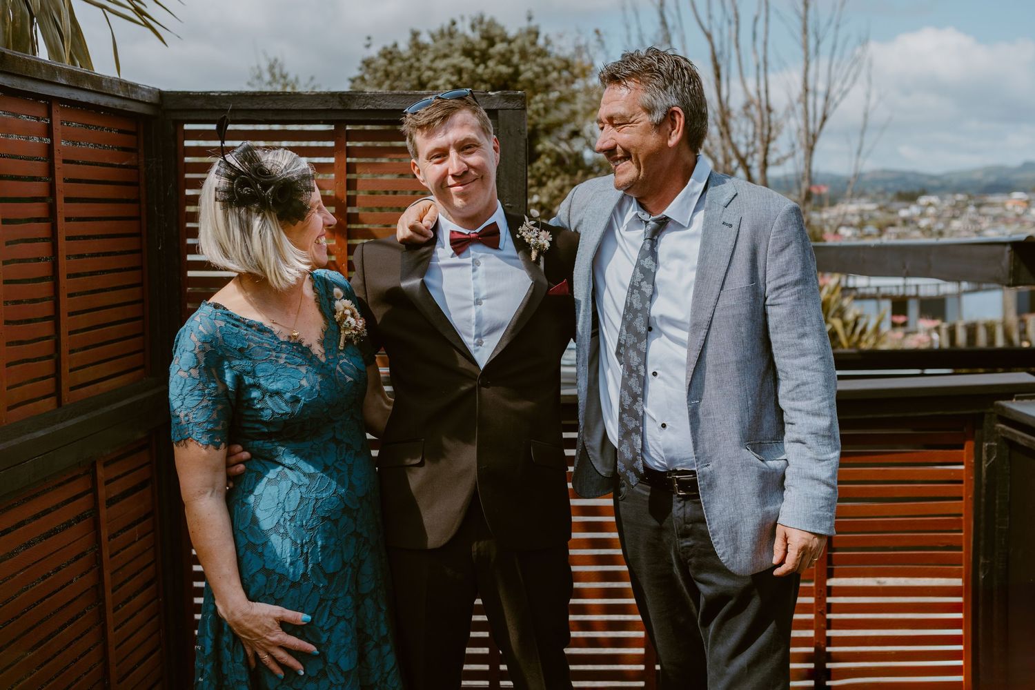 Three people sharing joyful moment during wedding celebration on wooden deck with scenic backdrop.