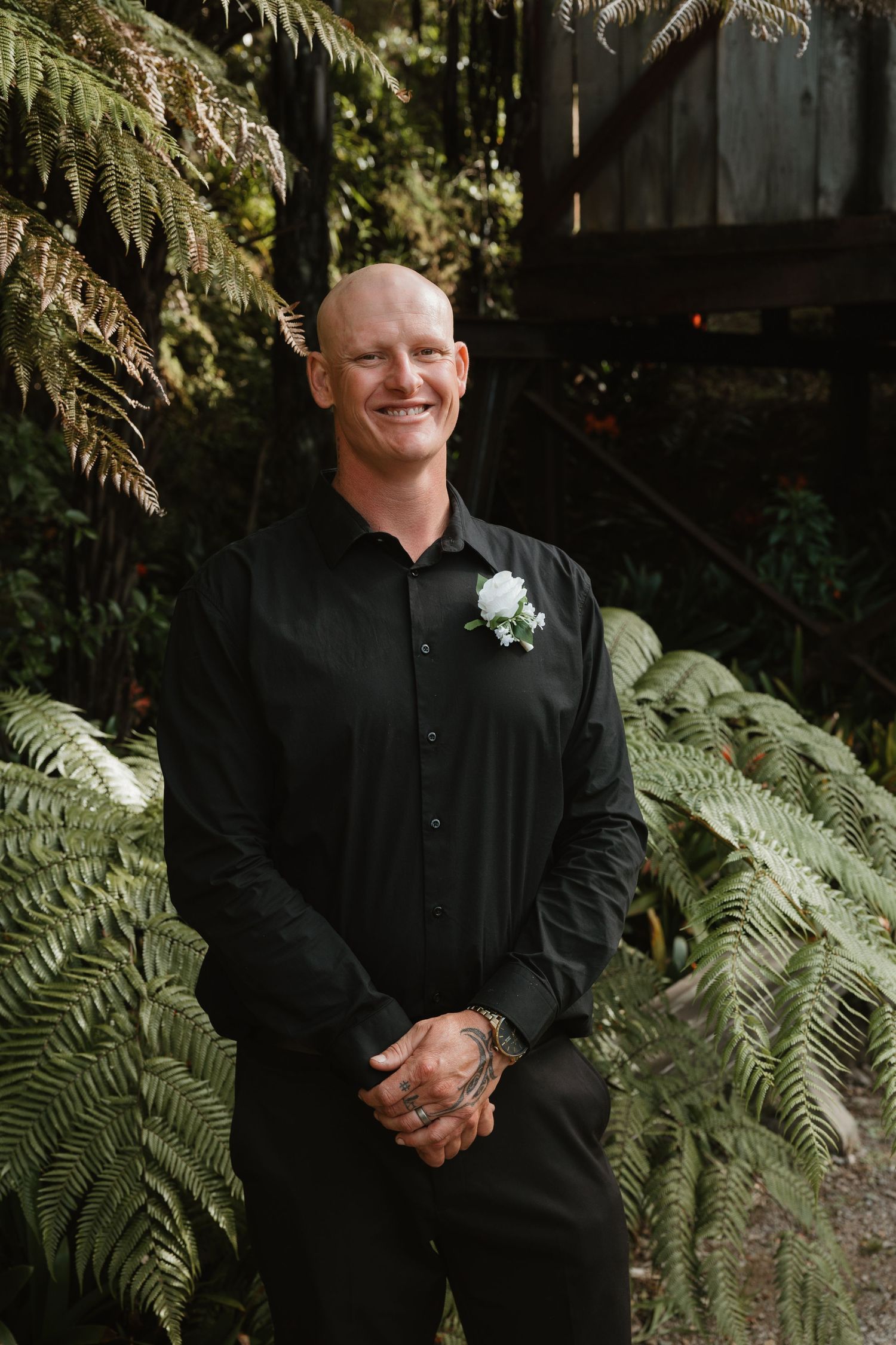 Person in black formal attire smiles while standing against lush green fern background in outdoor setting.