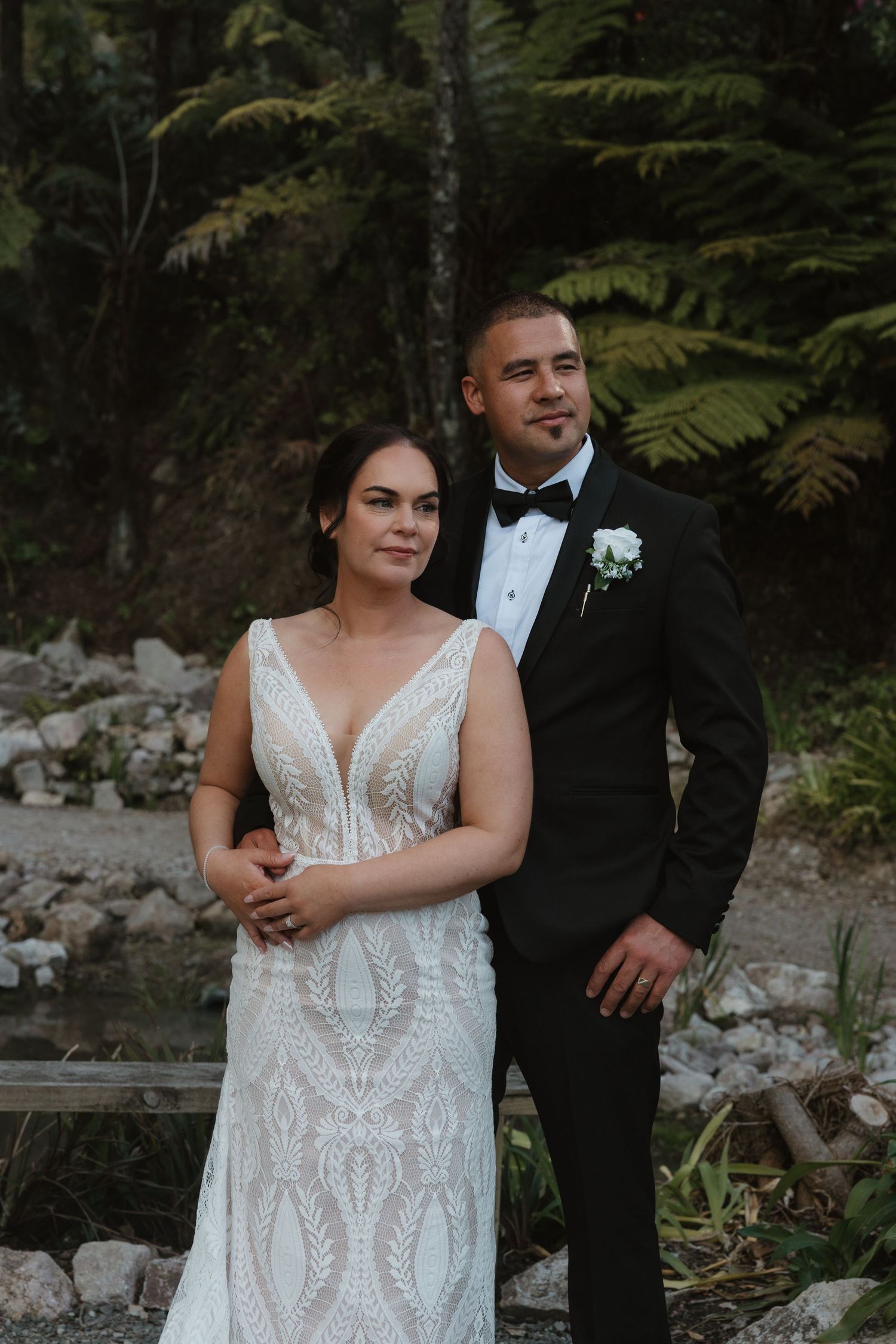 A couple poses together in formal wedding attire against a backdrop of lush ferns and forest greenery.