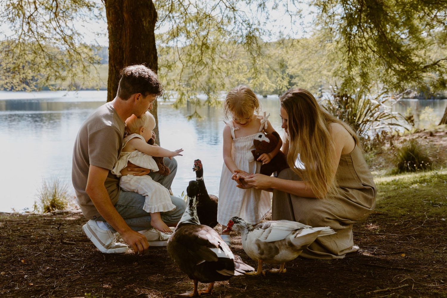 Family feeding ducks by a tranquil lake surrounded by lush trees on a sunny day. Mclaren Falls Photoshoot Tauranga