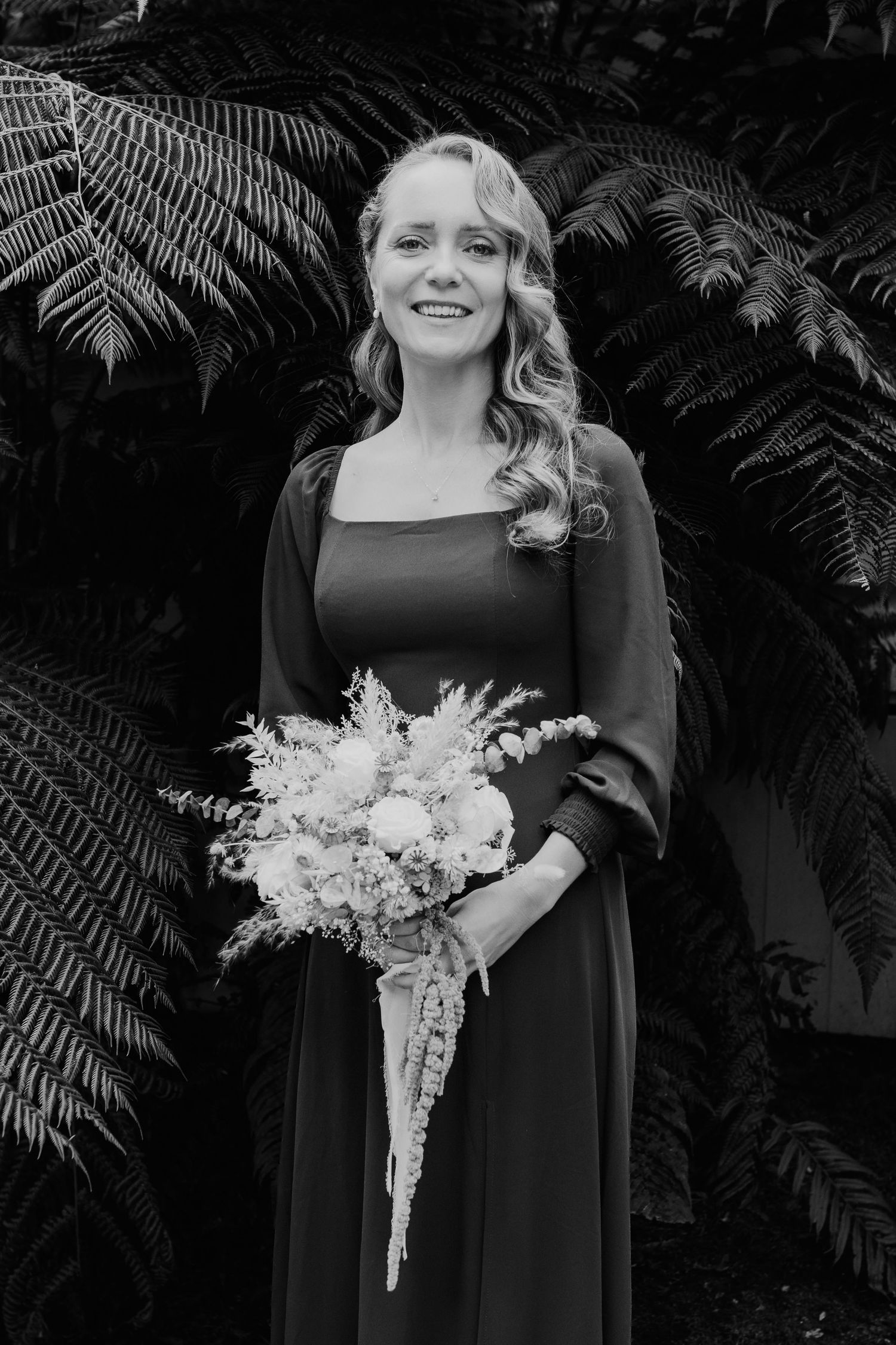 Black and white photo of a smiling figure in a long-sleeved dress holding a delicate white floral bouquet against fern foliage.