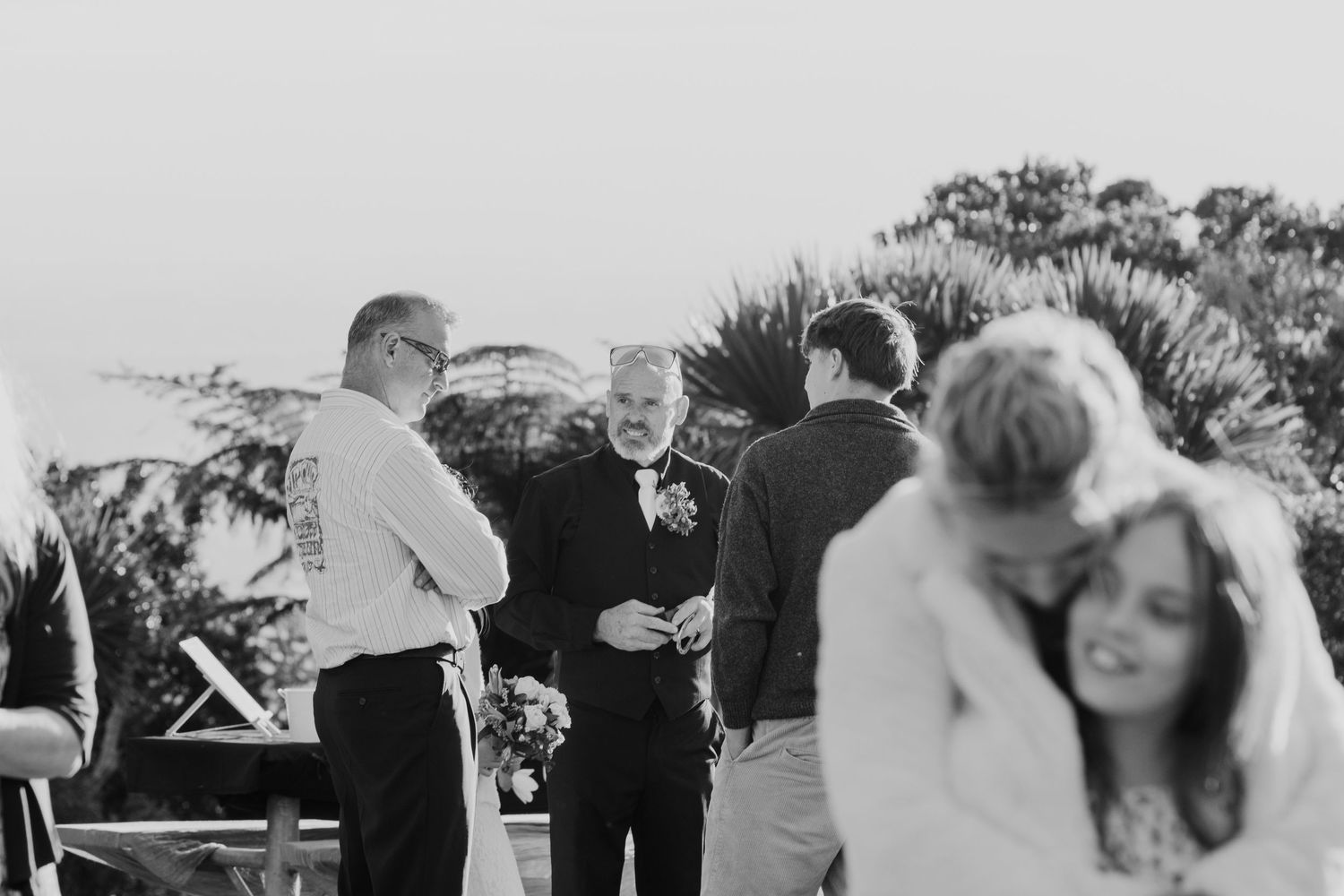 A black and white photo of guests mingling at an outdoor wedding reception with palm trees in the background.