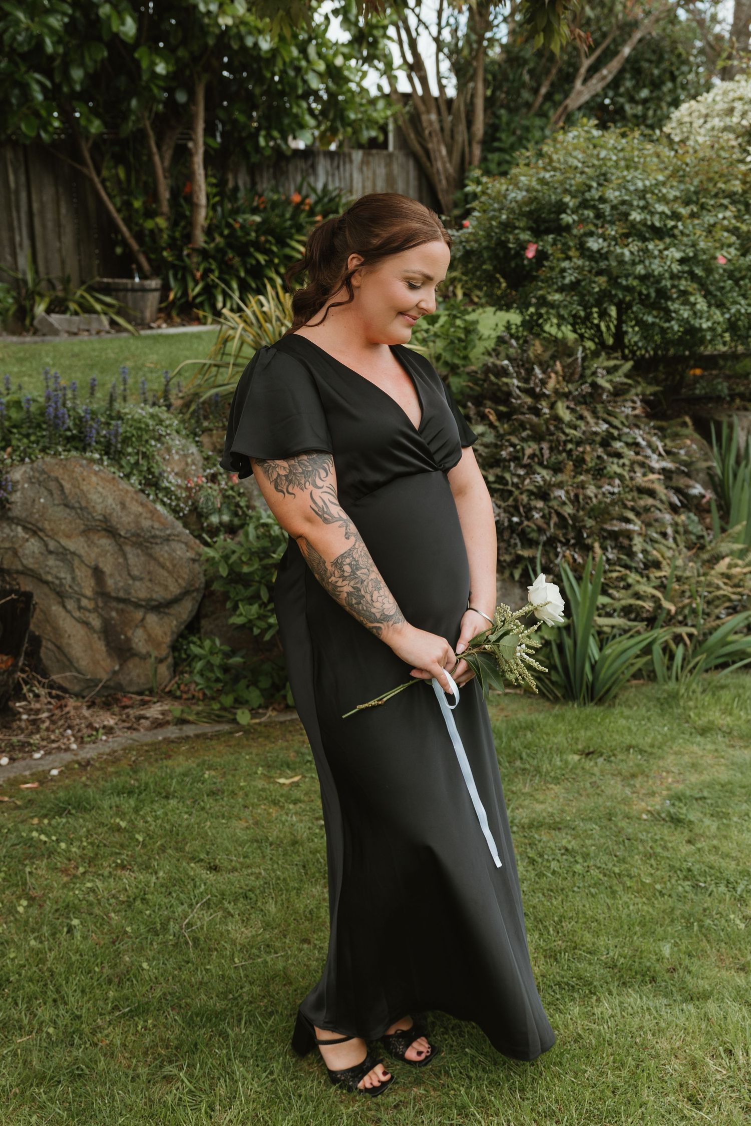 Person in black outfit kneeling on grass lawn with gardening tools in a landscaped backyard setting.