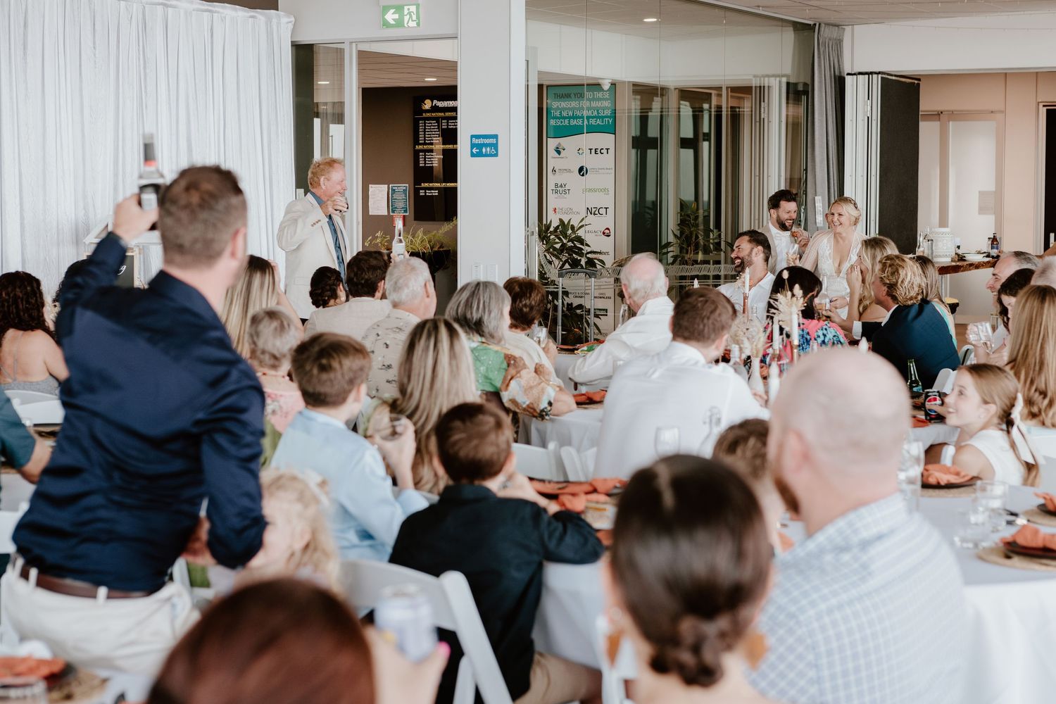 Wedding reception venue with white decor and guests seated at long tables under modern lighting. Papamoa Surf Life Saving Club