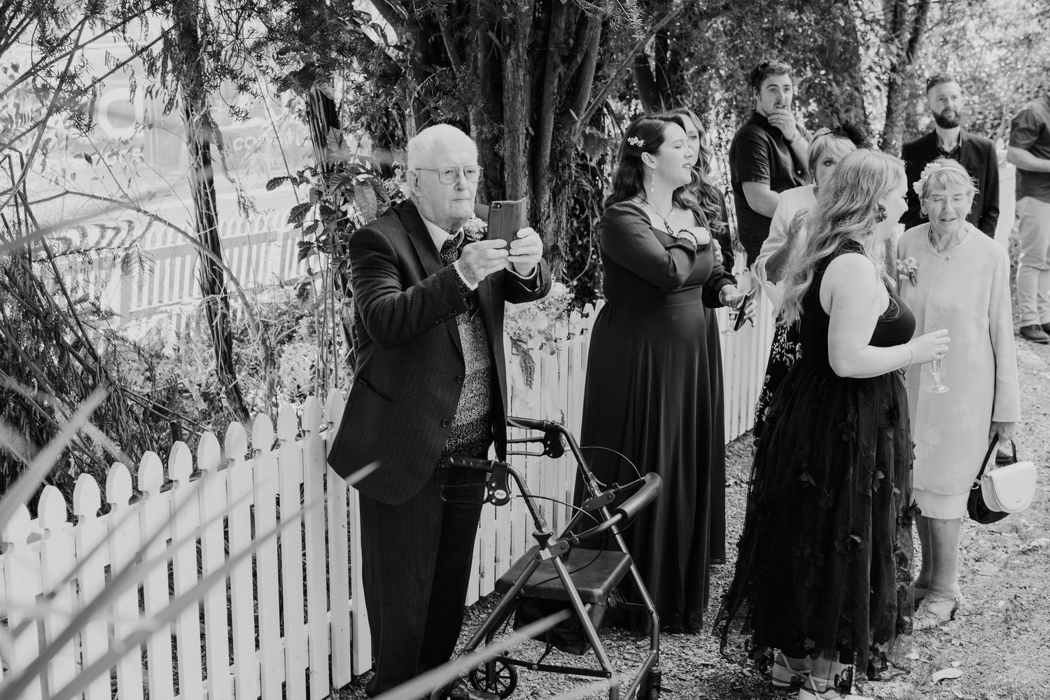 Wedding guests in formal attire gathered around a white picket fence.