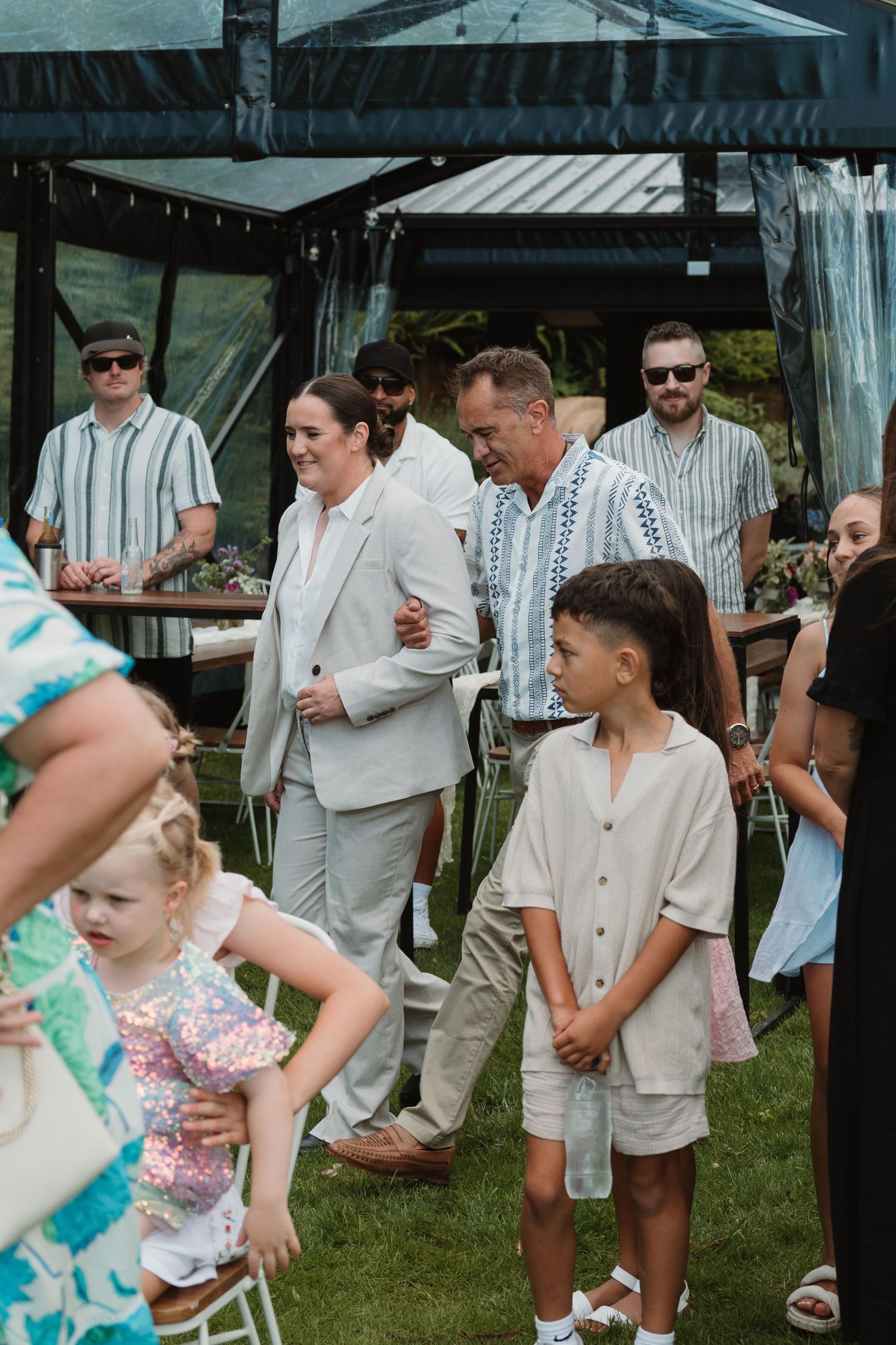 Guests in elegant attire gather outdoors at a summer wedding reception under a tent on green lawn.