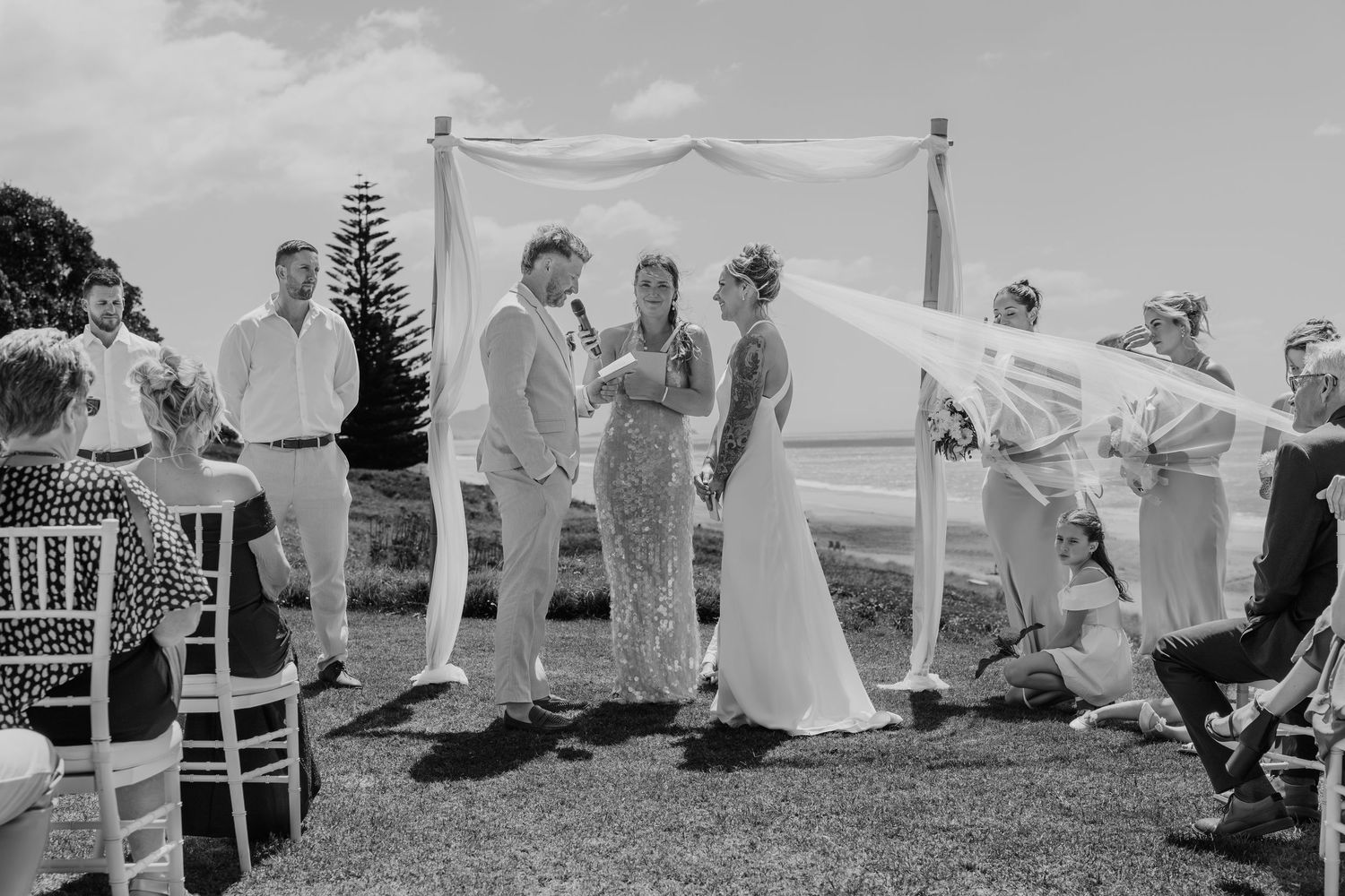 Black and white outdoor wedding ceremony with couple at altar, guests seated on white chairs, fabric canopy blowing in breeze.