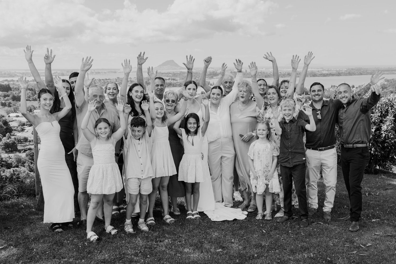 Black and white group photo of wedding party with raised hands celebrating outdoors with scenic view.