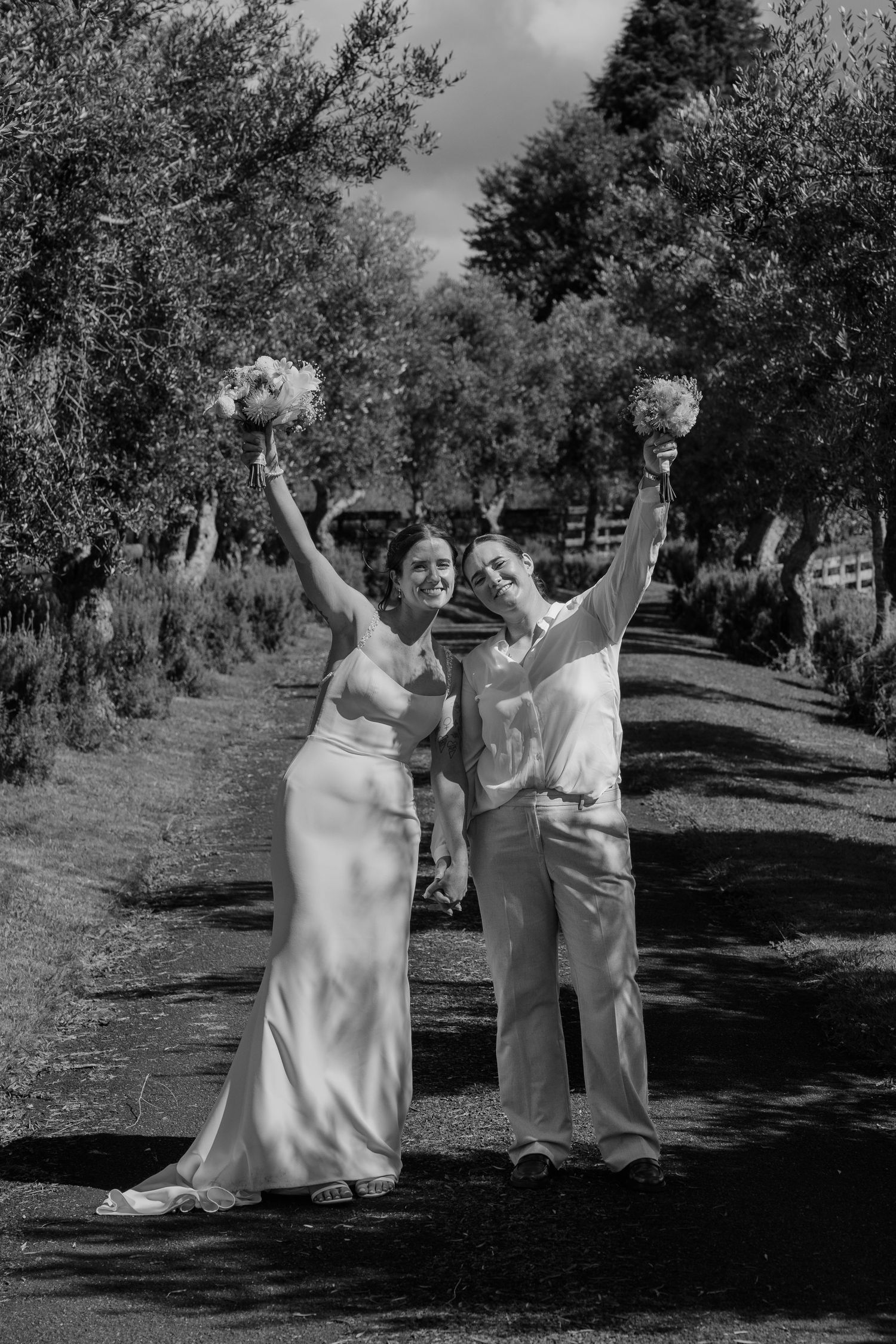 Black and white photo of two brides celebrating on a tree-lined path, raising bouquets joyfully on their wedding day.