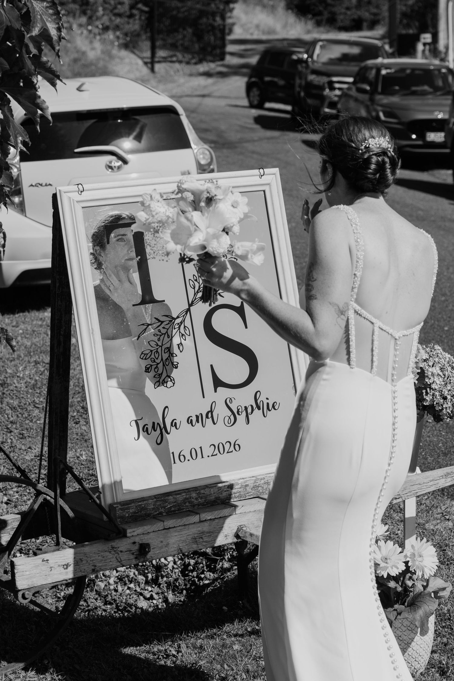 Bride in white gown admires wedding welcome sign for Tayla and Sophie dated 16.01.2026 outdoors.
