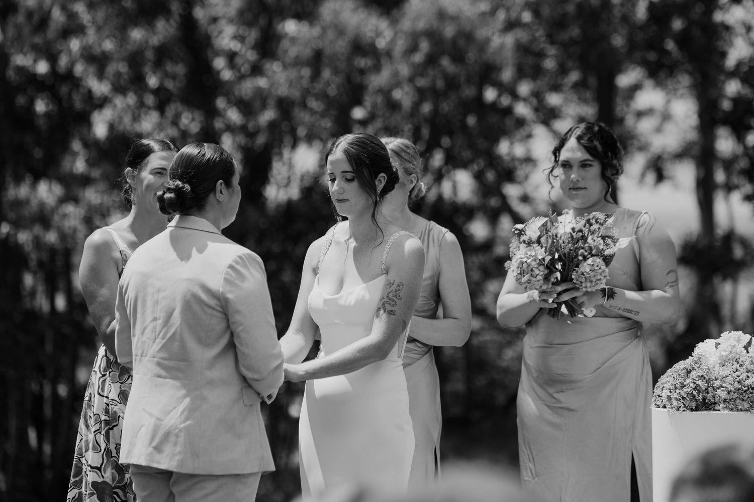 Black and white wedding photo of bride and groom holding hands with bridesmaids during outdoor ceremony.