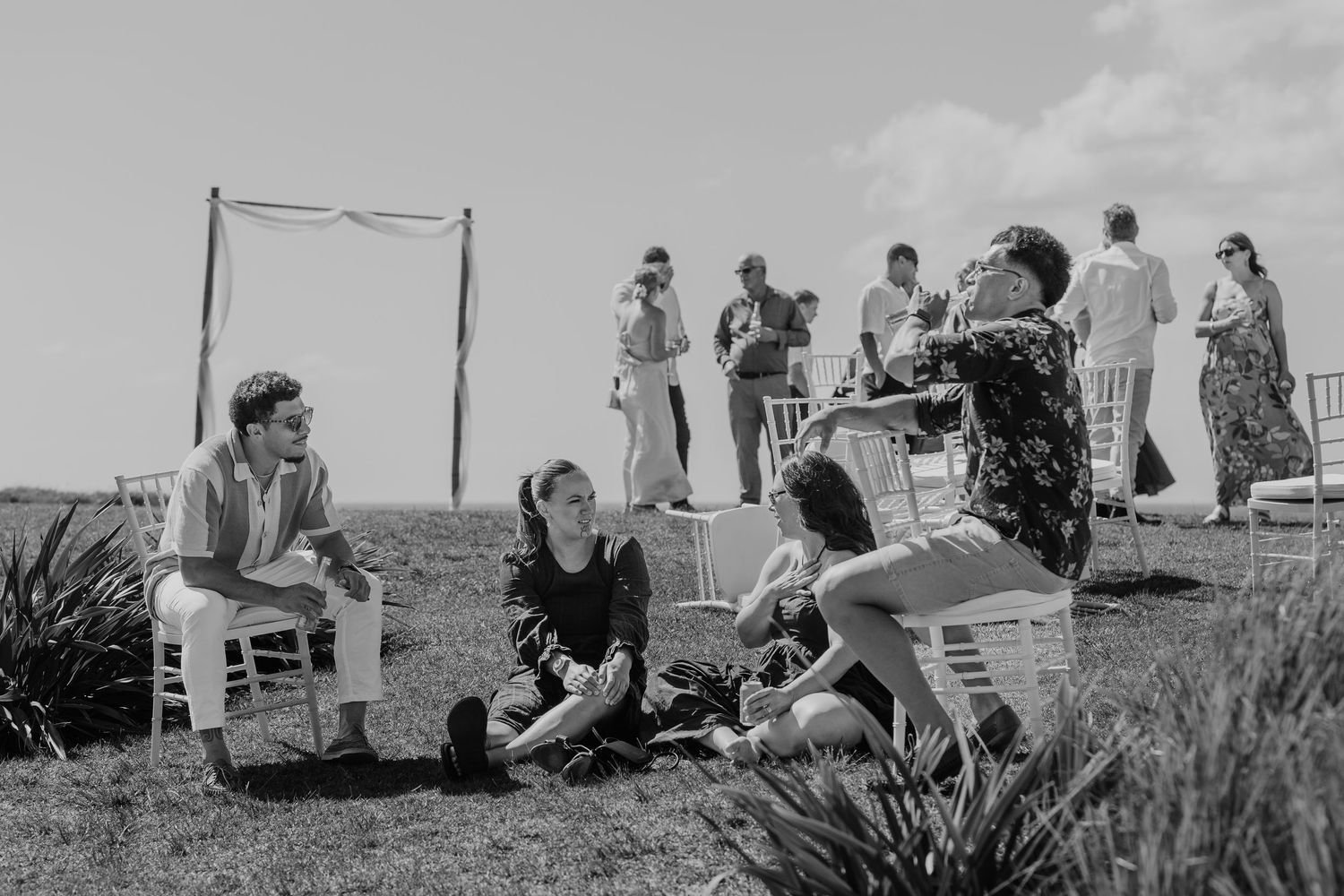 Black and white photo of guests relaxing on grass at an outdoor wedding ceremony on a sunny day.
