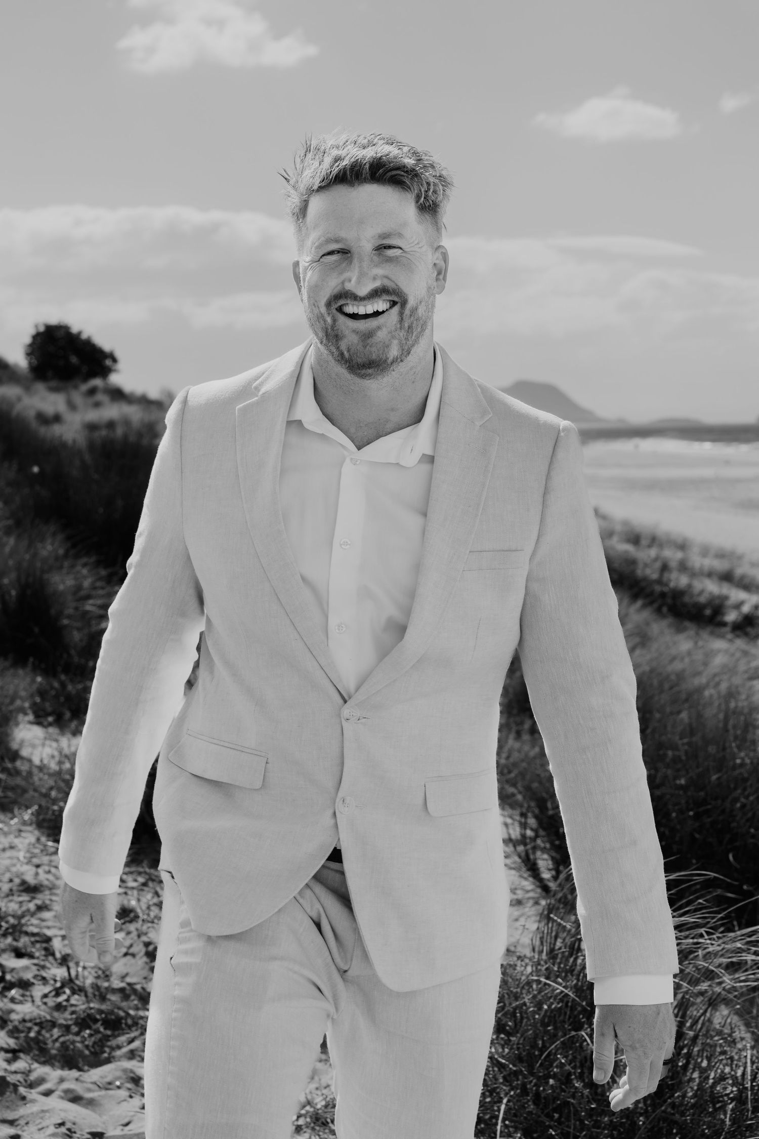 Smiling man in light suit posing on sandy beach with coastal scenery in black and white photography.