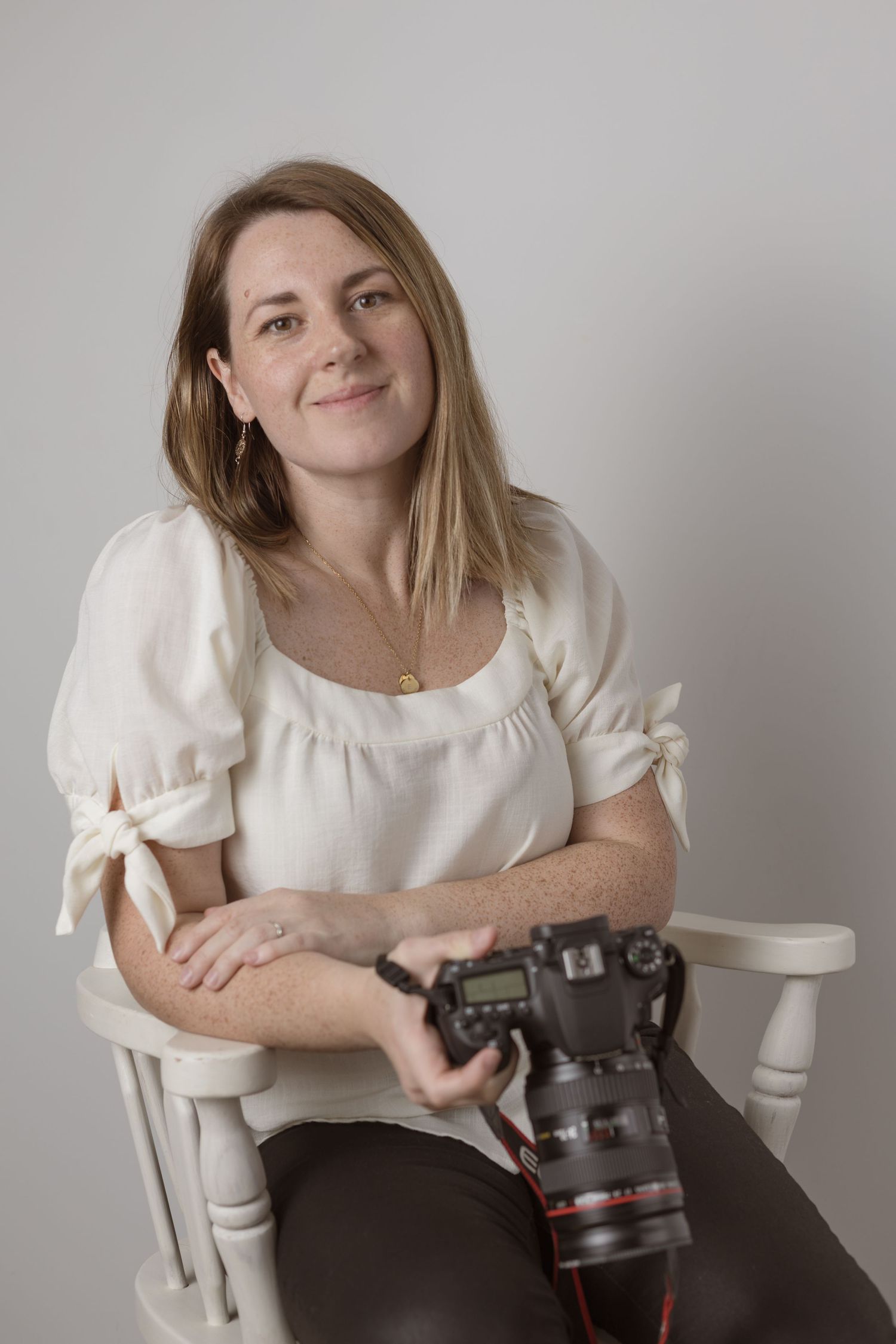Photographer in white blouse poses with professional camera in studio against neutral background.