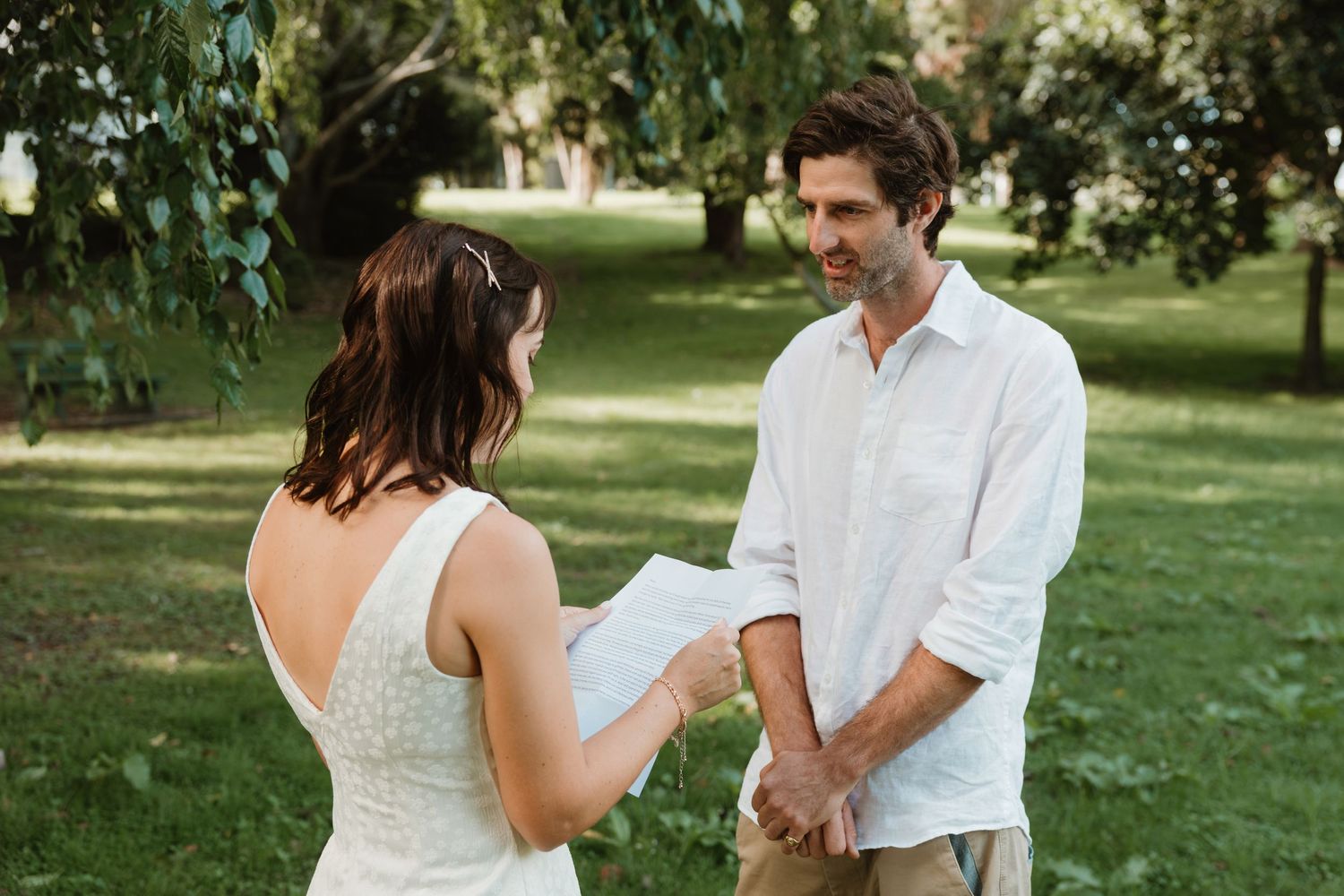Couple exchanging wedding vows outdoors in a lush green garden, bride in white dress holding paper vows.