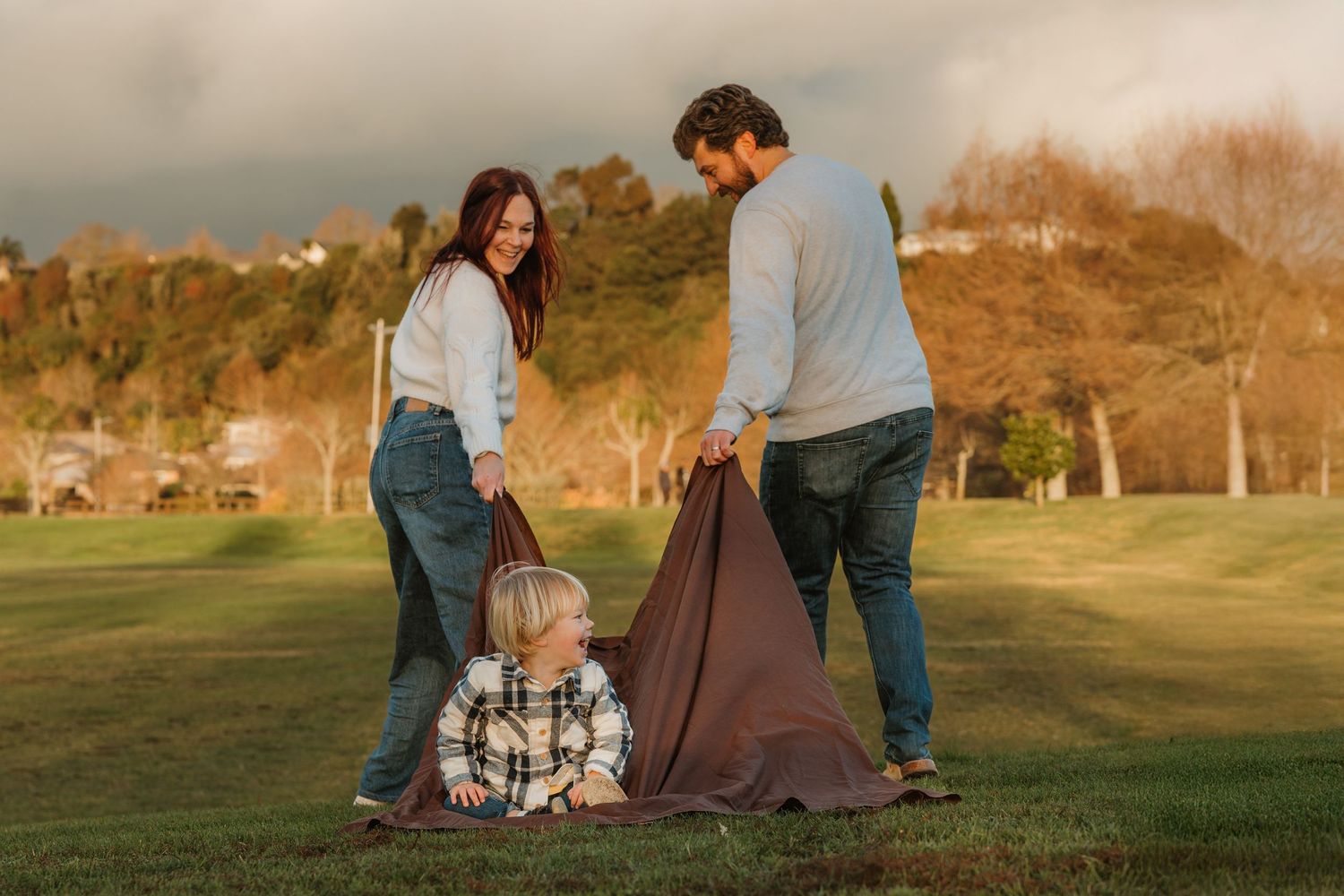 A young family plays with a brown blanket on a green lawn at golden hour, with a toddler sitting in the grass.