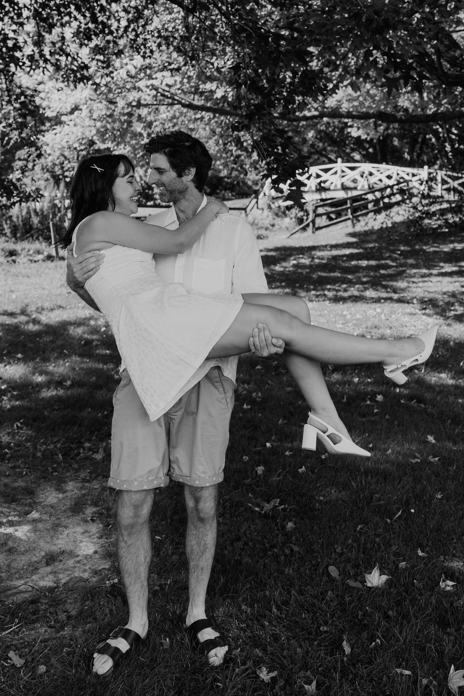 Black and white photo of a man carrying a woman in his arms outdoors, both smiling and dressed in white summer outfits.