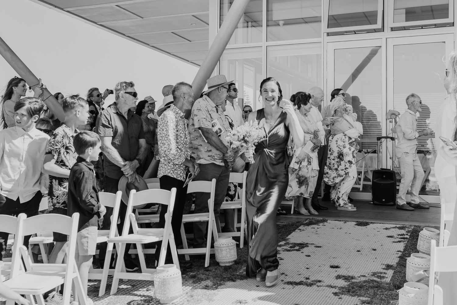 Black and white photo of wedding guests seated in white chairs at an outdoor ceremony. Papamoa Surf Lifesaving Club Wedding