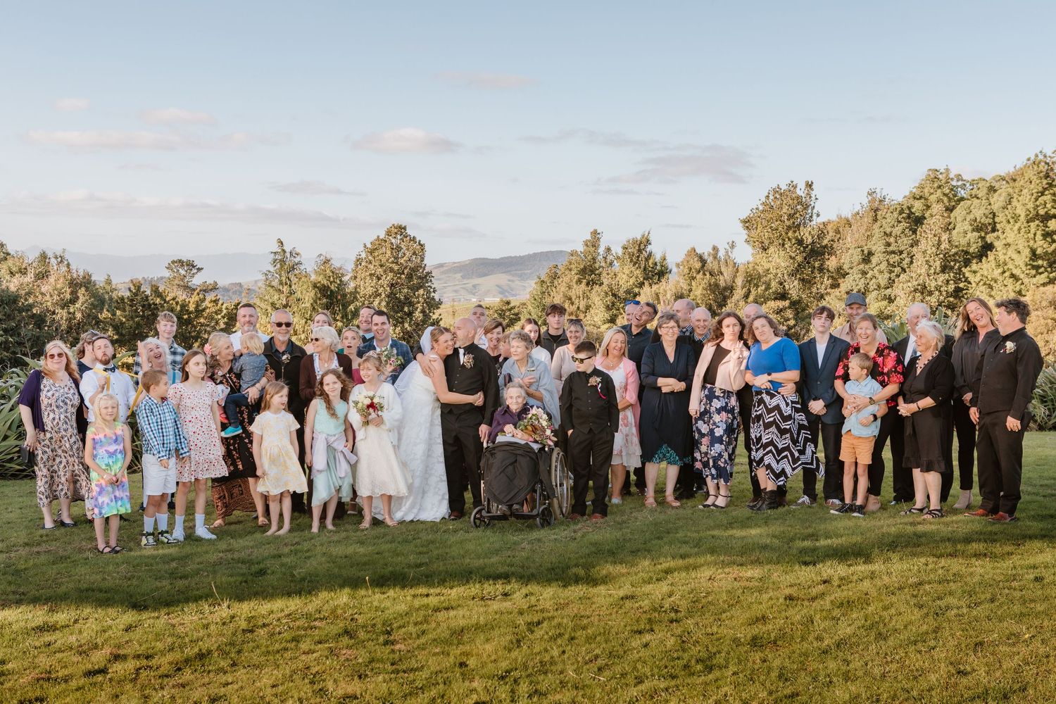A large group gathers on a grassy hilltop for an outdoor wedding ceremony at golden hour.
